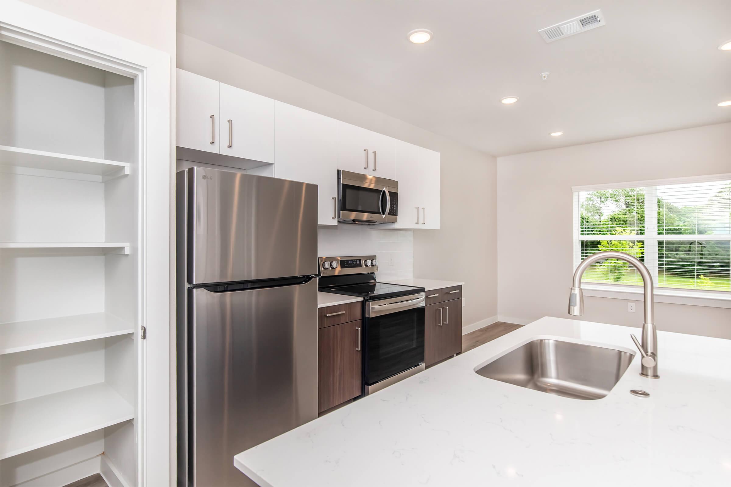 Modern kitchen with stainless steel appliances including a refrigerator, oven, and microwave. Features white cabinetry, a large sink, and a spacious countertop. There's a built-in pantry on the left and a window providing natural light and a view of greenery outside. Bright, clean, and contemporary design.