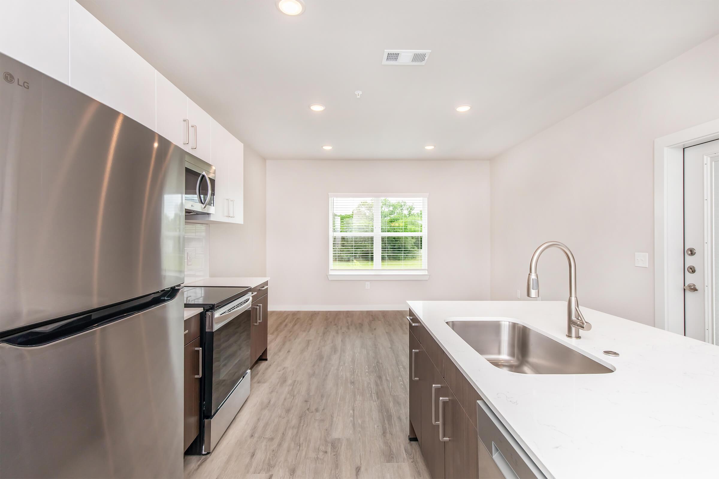 Modern kitchen featuring stainless steel appliances, including a refrigerator and microwave, sleek cabinetry, and a large island with a sink. The space is well-lit with recessed lighting and a window providing natural light, complemented by light-colored walls and wooden flooring.