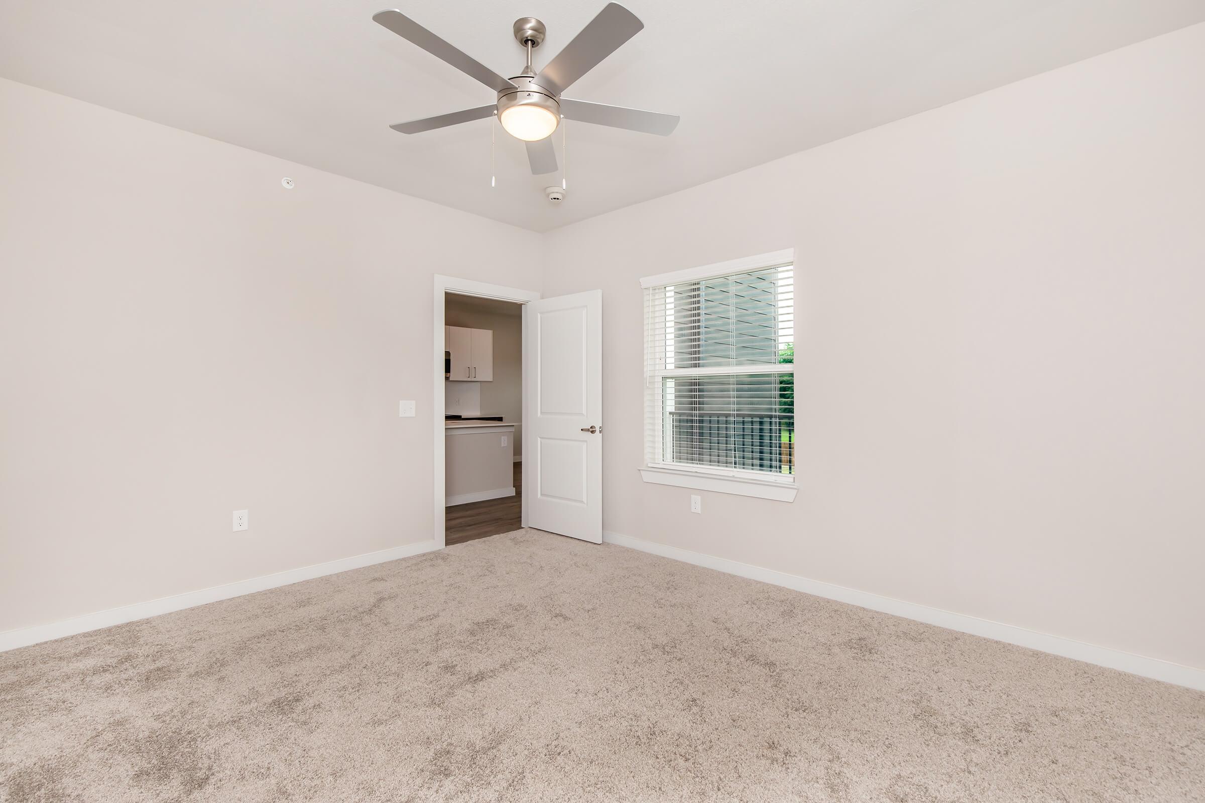 A spacious, light neutral-colored room with carpeted flooring, a ceiling fan, and a window allowing natural light. A doorway leads to another room with a view of a kitchen area. The walls are painted in a soft hue, creating a calm atmosphere. Ideal for a cozy living or bedroom space.