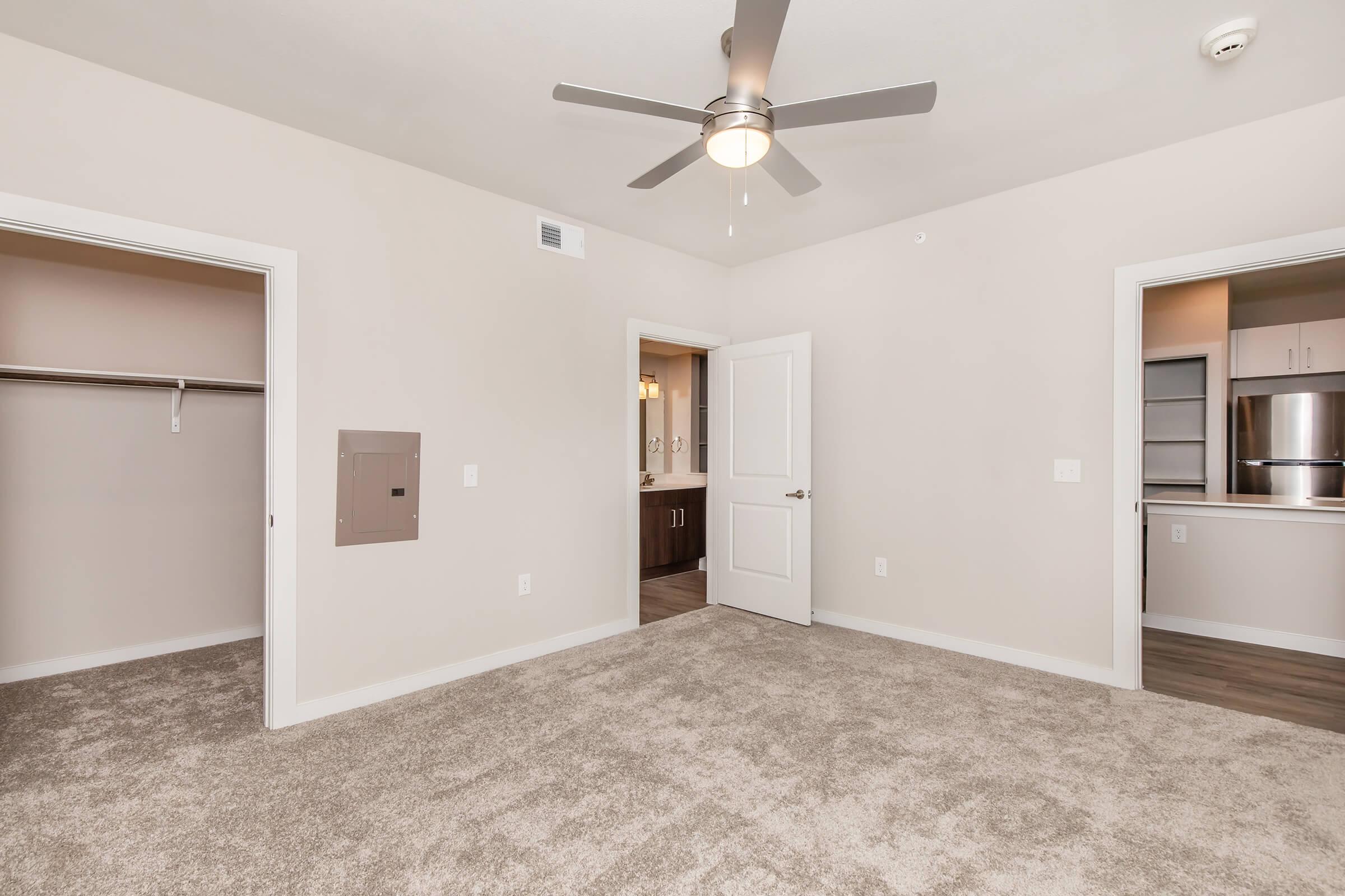 A view of a spacious room with light beige walls and soft, carpeted flooring. There are two doorways leading to other areas, one with a closet. A ceiling fan is mounted overhead, and there is a wall-mounted intercom system. A glimpse of an adjacent kitchen area is visible through an open doorway.
