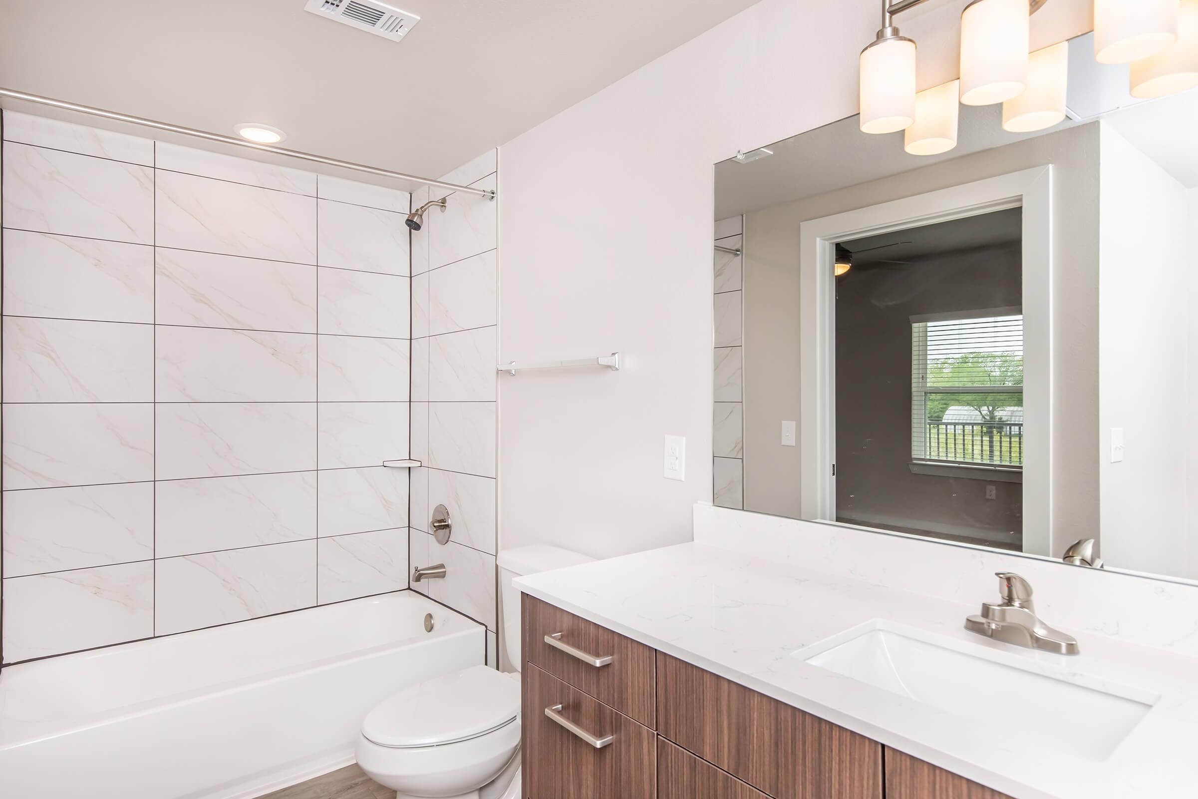 A modern bathroom featuring a white bathtub with a shower, marble-tiled walls, and a sleek countertop with a sink. The cabinetry is wooden, and there is a large mirror above the sink. Soft lighting from pendant fixtures illuminates the space. A window provides natural light and a view of the outdoors.
