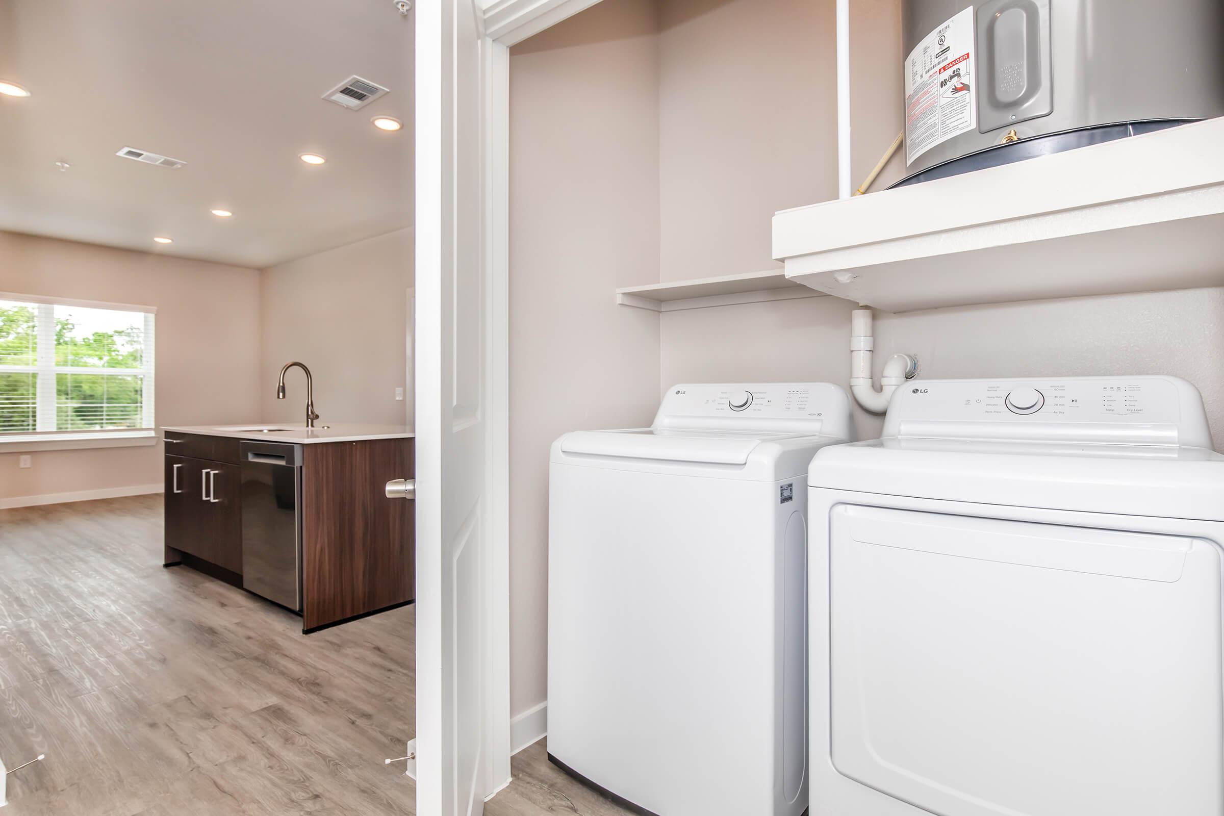 A laundry area featuring stacked washing machine and dryer units beside a modern kitchen with a sink and cabinets. Natural light filters in through a window, illuminating the space with light-colored walls and flooring.