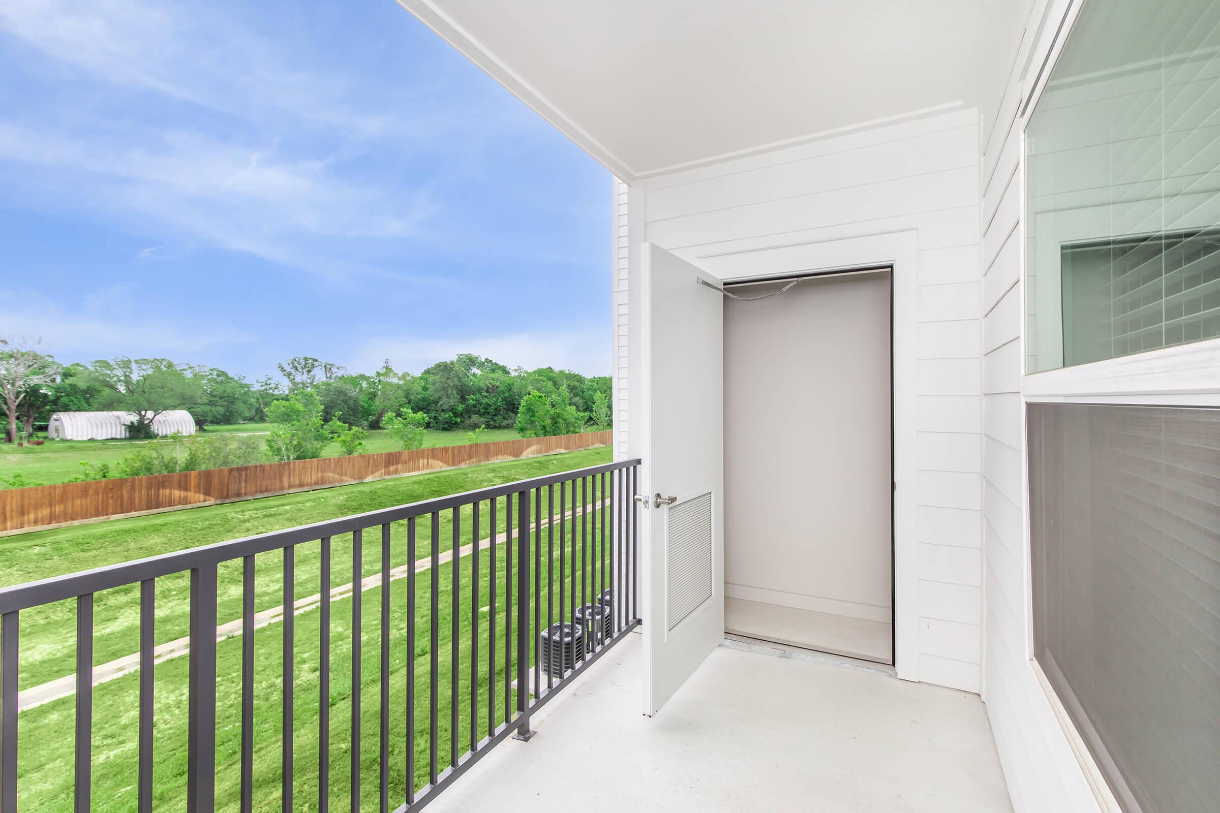 A view from a modern balcony showcasing an open door leading inside. The balcony features a sleek railing, and the landscape beyond includes a well-maintained green lawn bordered by trees and a wooden fence. The sky above is clear and bright, adding to the inviting atmosphere.