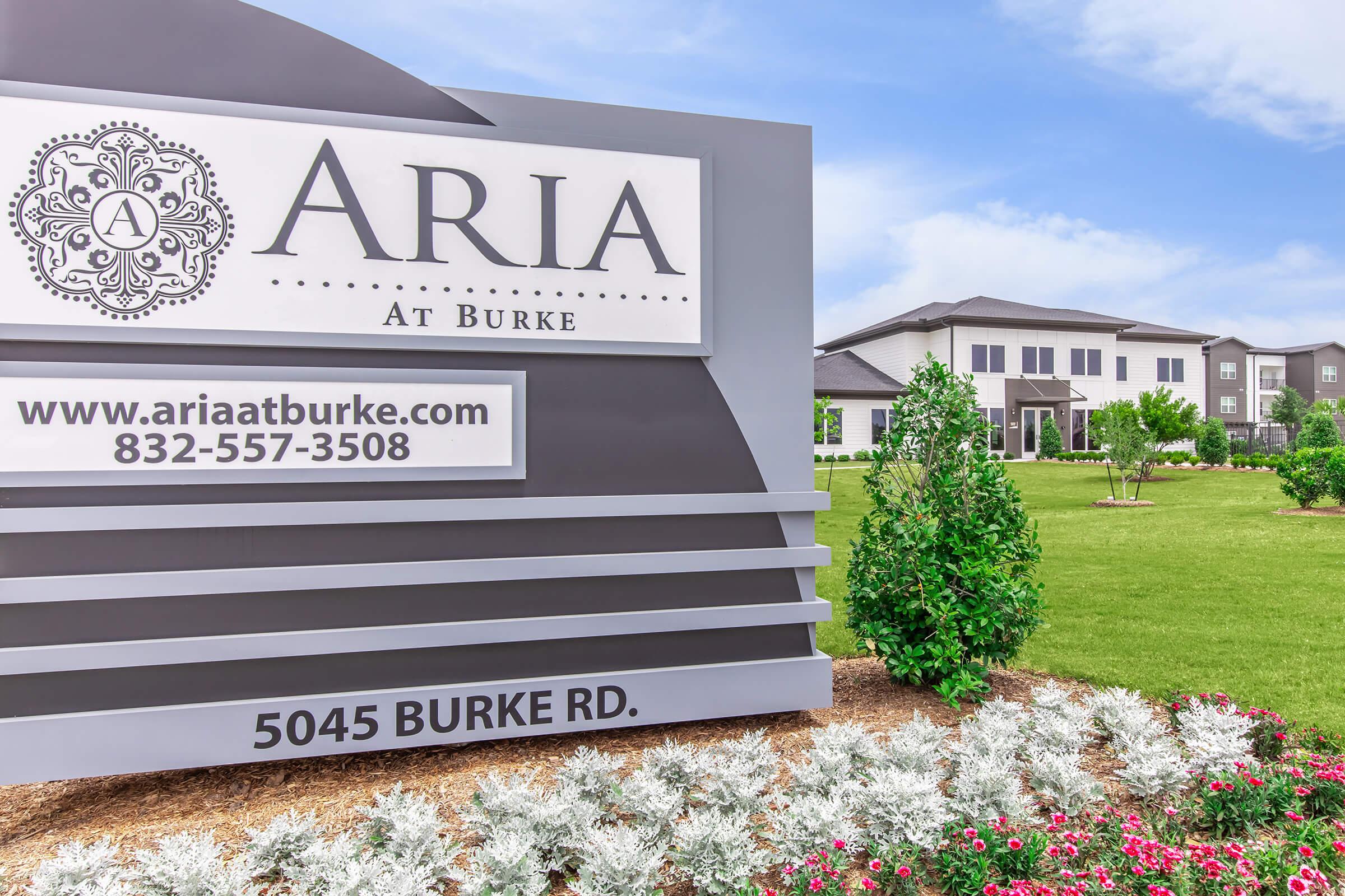 Sign for "Aria at Burke" with contact information, featuring a welcoming landscape of greenery and flowering plants in front of modern residential buildings. The sign includes the address 5045 Burke Rd and has a decorative emblem at the top. Clear blue sky in the background enhances the inviting atmosphere.