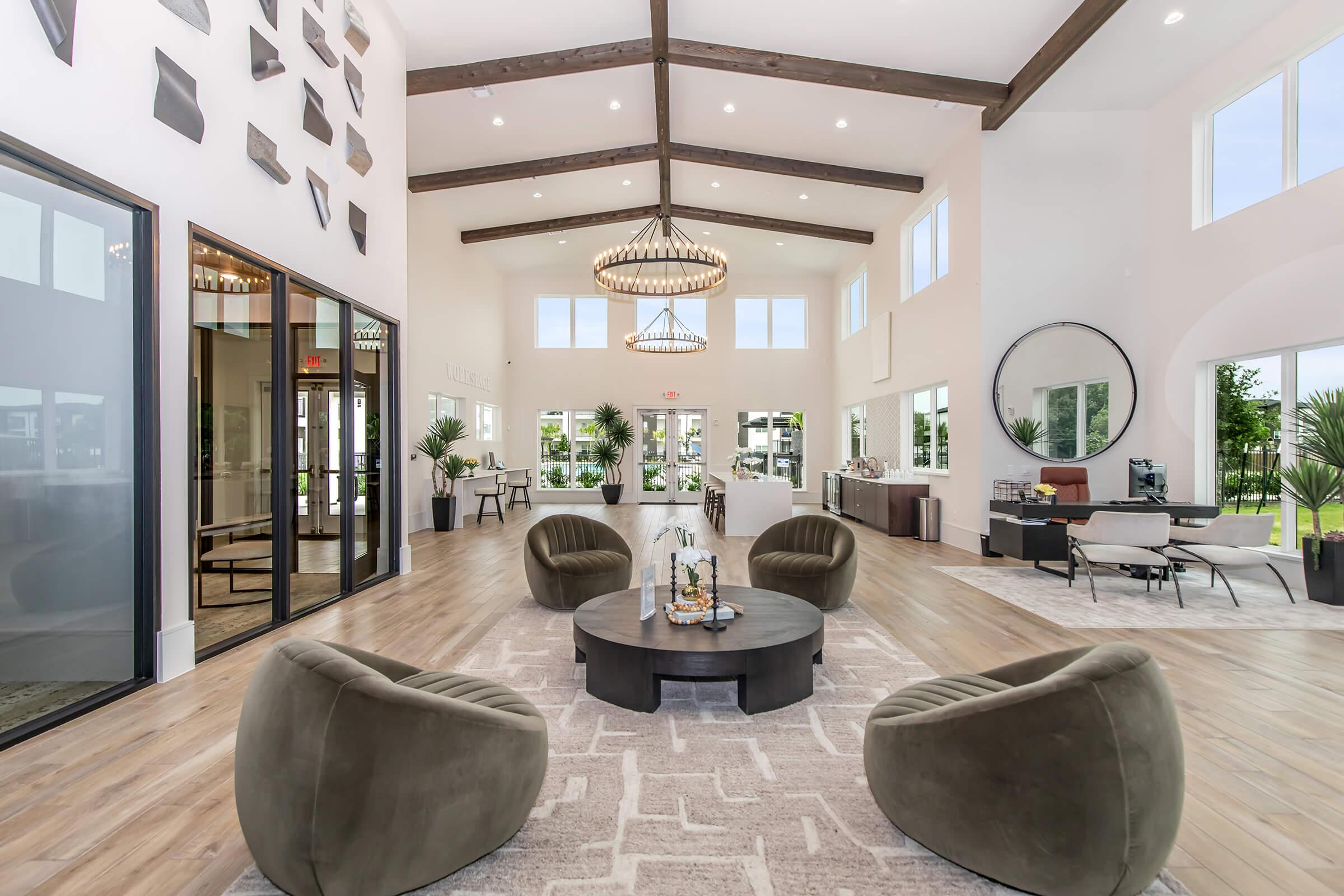 A spacious, modern lobby area featuring high ceilings with wooden beams. Four round, green chairs are arranged around a central coffee table. Large windows allow natural light to fill the space, and stylish decor, including plants and artwork, enhances the inviting atmosphere. A circular mirror and contemporary furnishings complete the design.