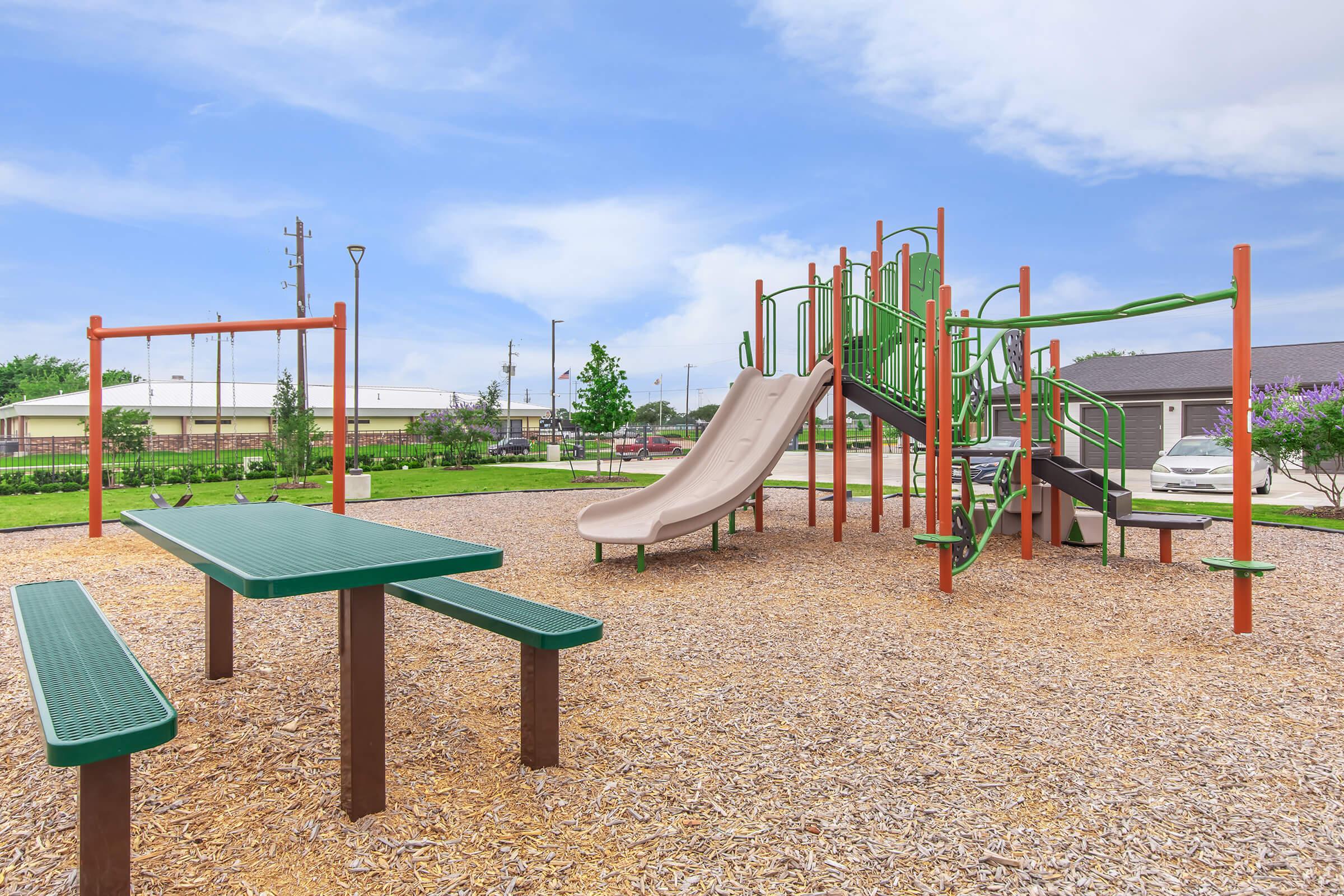 A colorful playground featuring green and tan slides, climbing structures, and picnic tables surrounded by mulch and grass. The sky is bright with clouds, and there are trees in the background. Suitable for children's outdoor play and recreational activities.