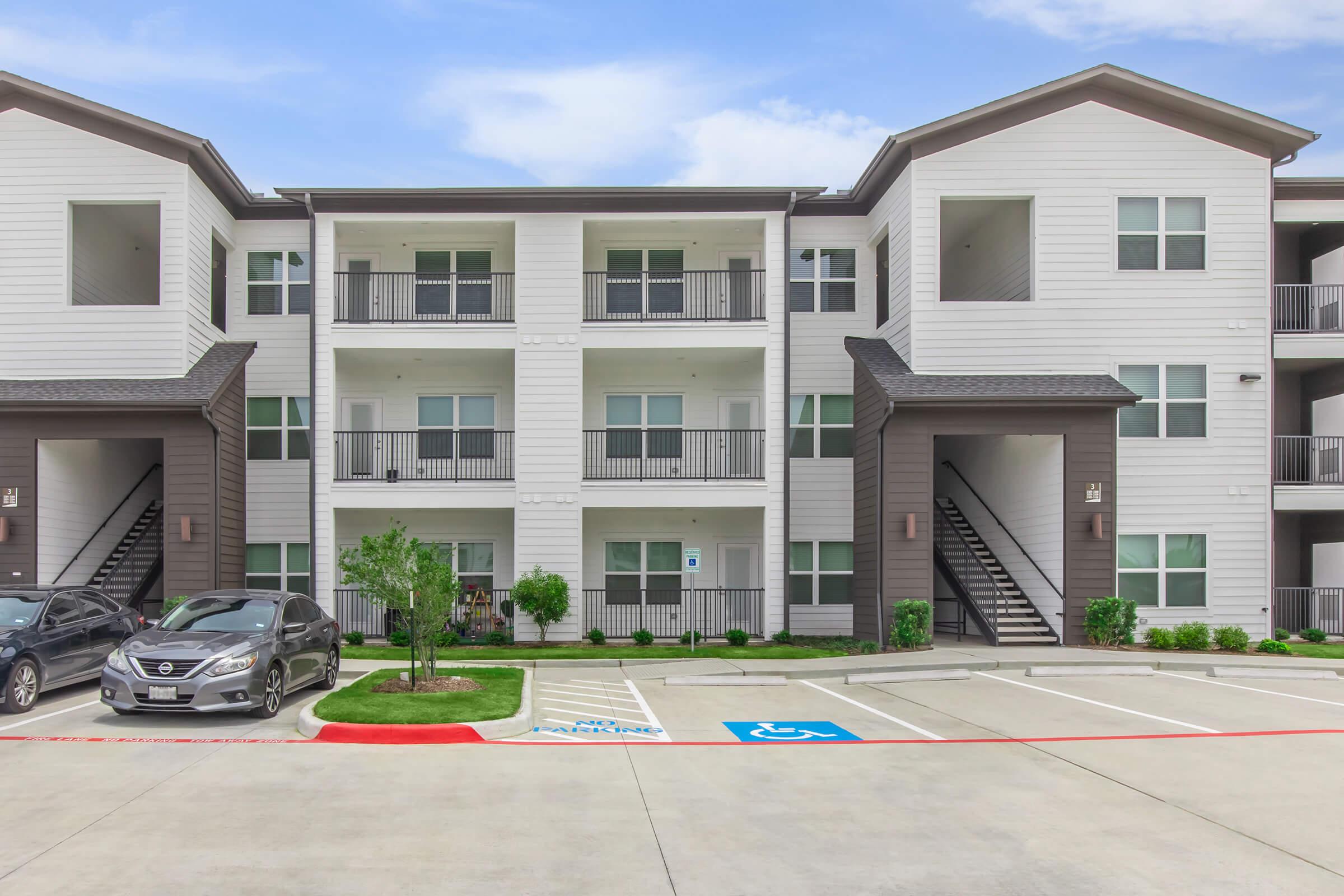 Modern apartment building featuring three stories, with balconies on the upper floors. The exterior is a combination of light and dark siding. Parking spaces are available in front, including designated spots. The area is landscaped with small shrubs and greenery. Cloudy sky in the background.