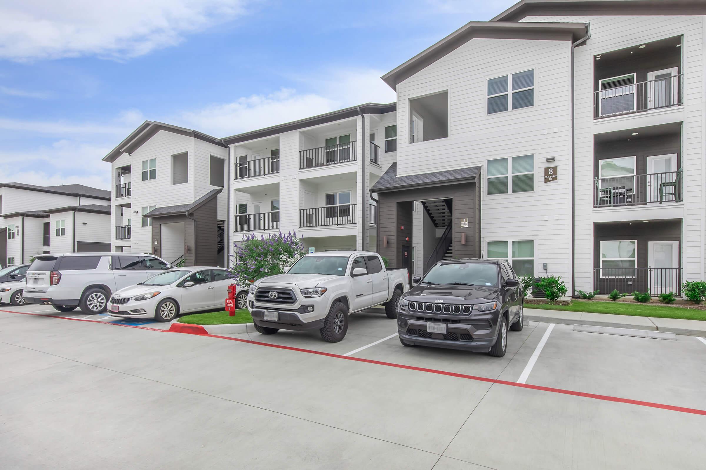 A modern apartment complex featuring multiple three-story buildings with white and dark exterior finishes. The foreground includes a parking lot with several parked vehicles, including a gray pickup truck and a black SUV. Lush landscaping and a clear blue sky are visible in the background.