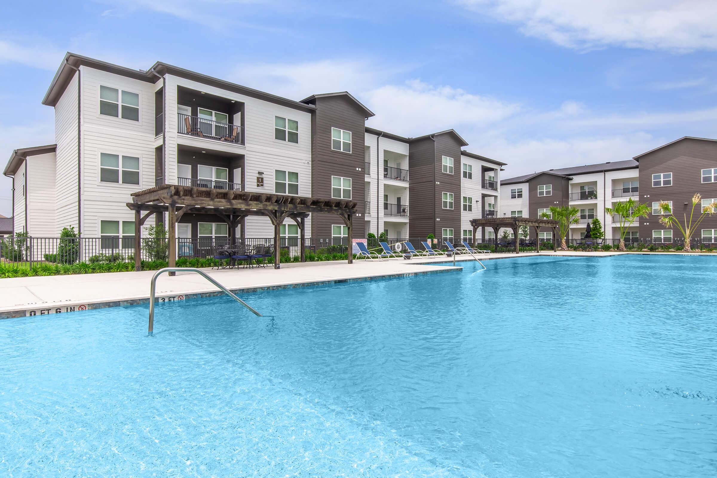 Swimming pool with a clear blue surface in the foreground, surrounded by a landscaped area. In the background, modern apartment buildings with balconies and shaded sitting areas are visible under a partly cloudy sky.