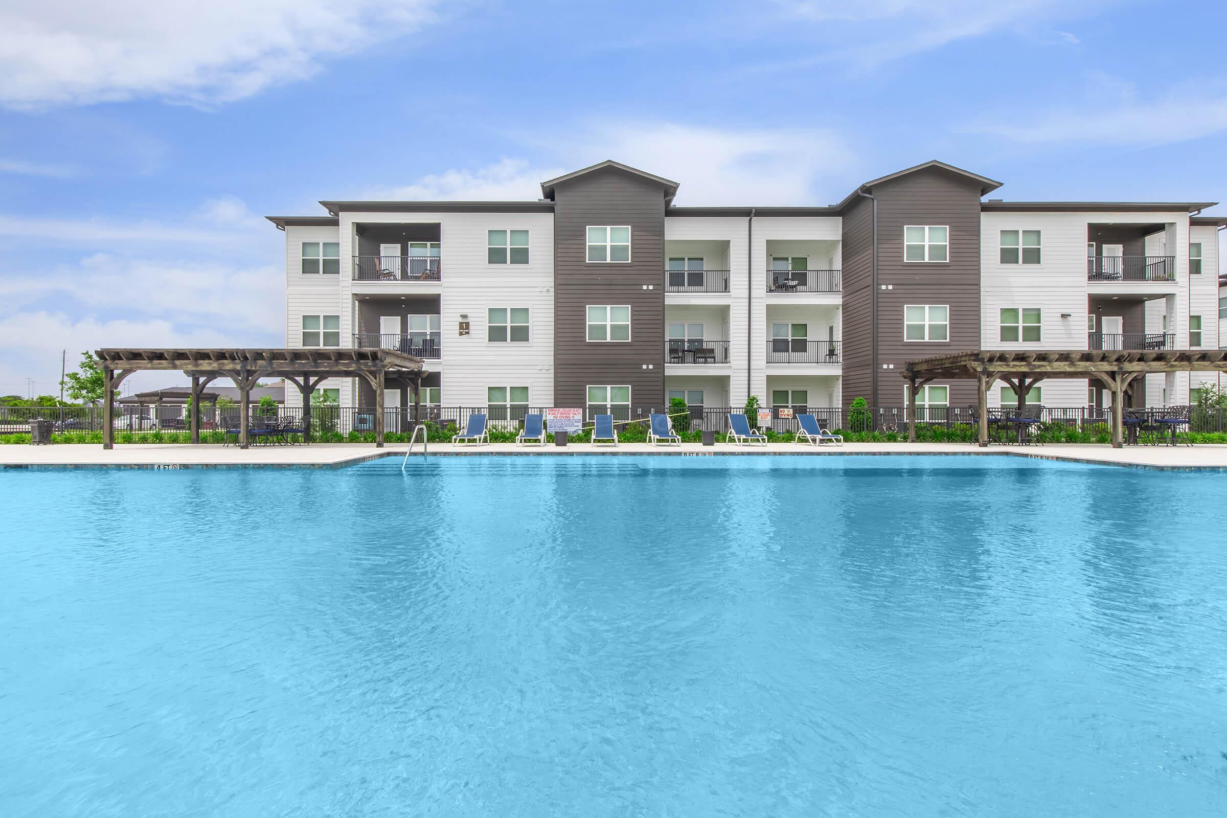 A modern apartment complex featuring multiple stories with balconies, situated by a large swimming pool. The pool area has lounge chairs and a shaded pergola, surrounded by green landscaping. The sky is clear with a few clouds, creating a bright and inviting atmosphere.