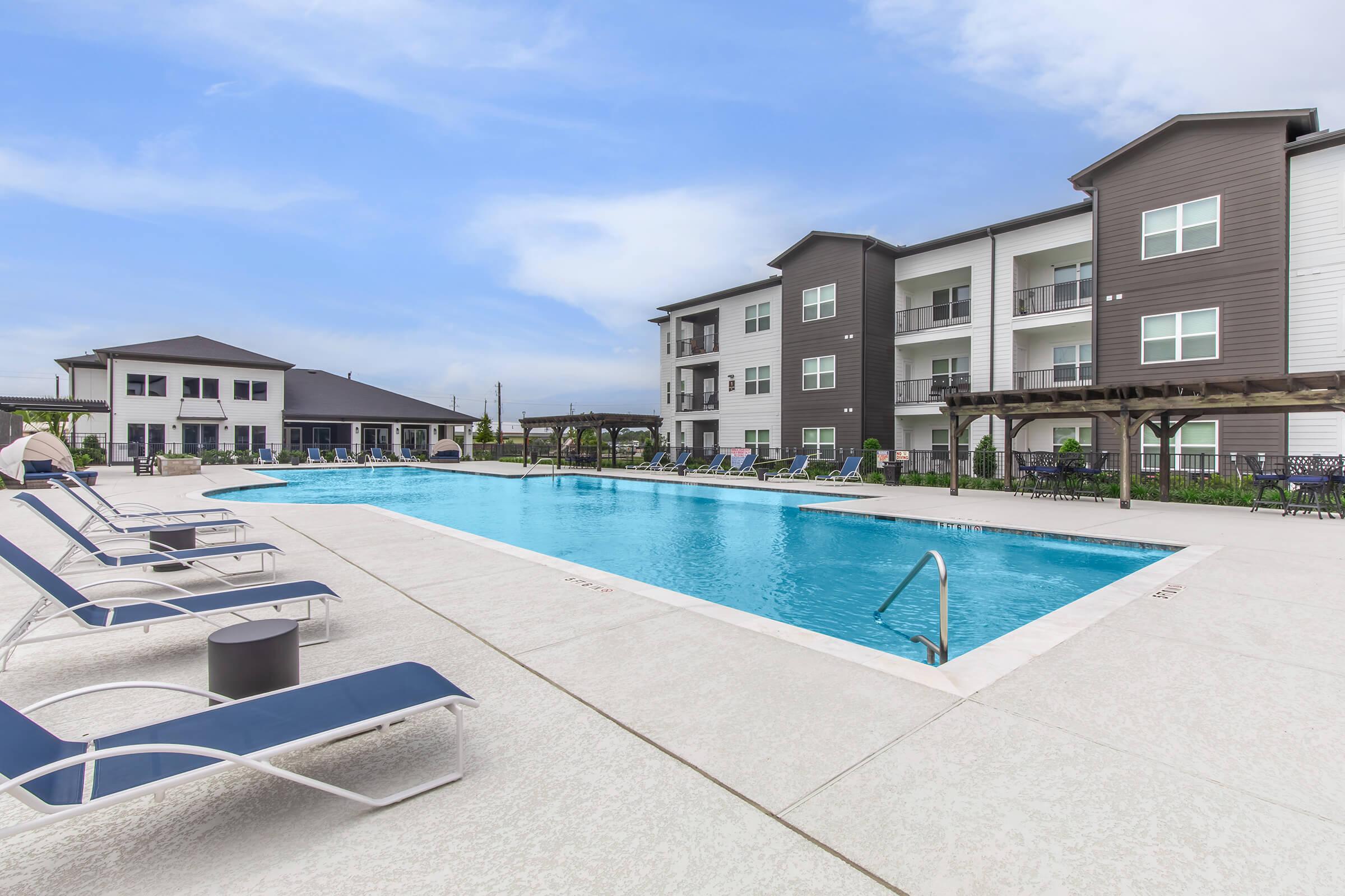 A spacious outdoor swimming pool surrounded by lounge chairs, with modern apartment buildings in the background. The area is well-maintained, featuring green landscaping and blue sky overhead, creating a relaxing atmosphere for residents and visitors.