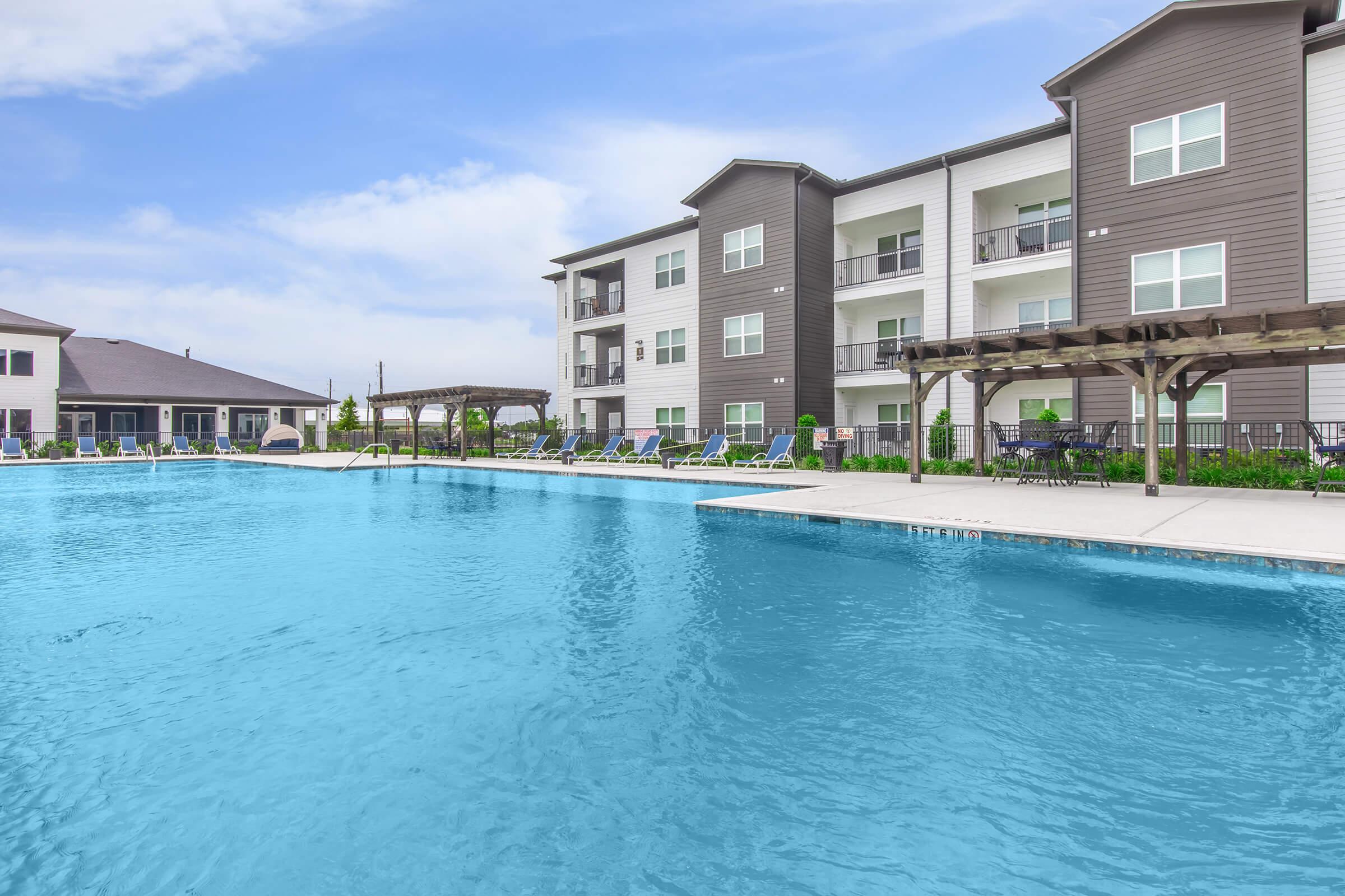 A spacious outdoor swimming pool surrounded by modern apartment buildings. The pool features clear blue water and is lined with lounge chairs. In the background, there are shaded pergolas and additional seating areas, all set against a bright blue sky with a few clouds.