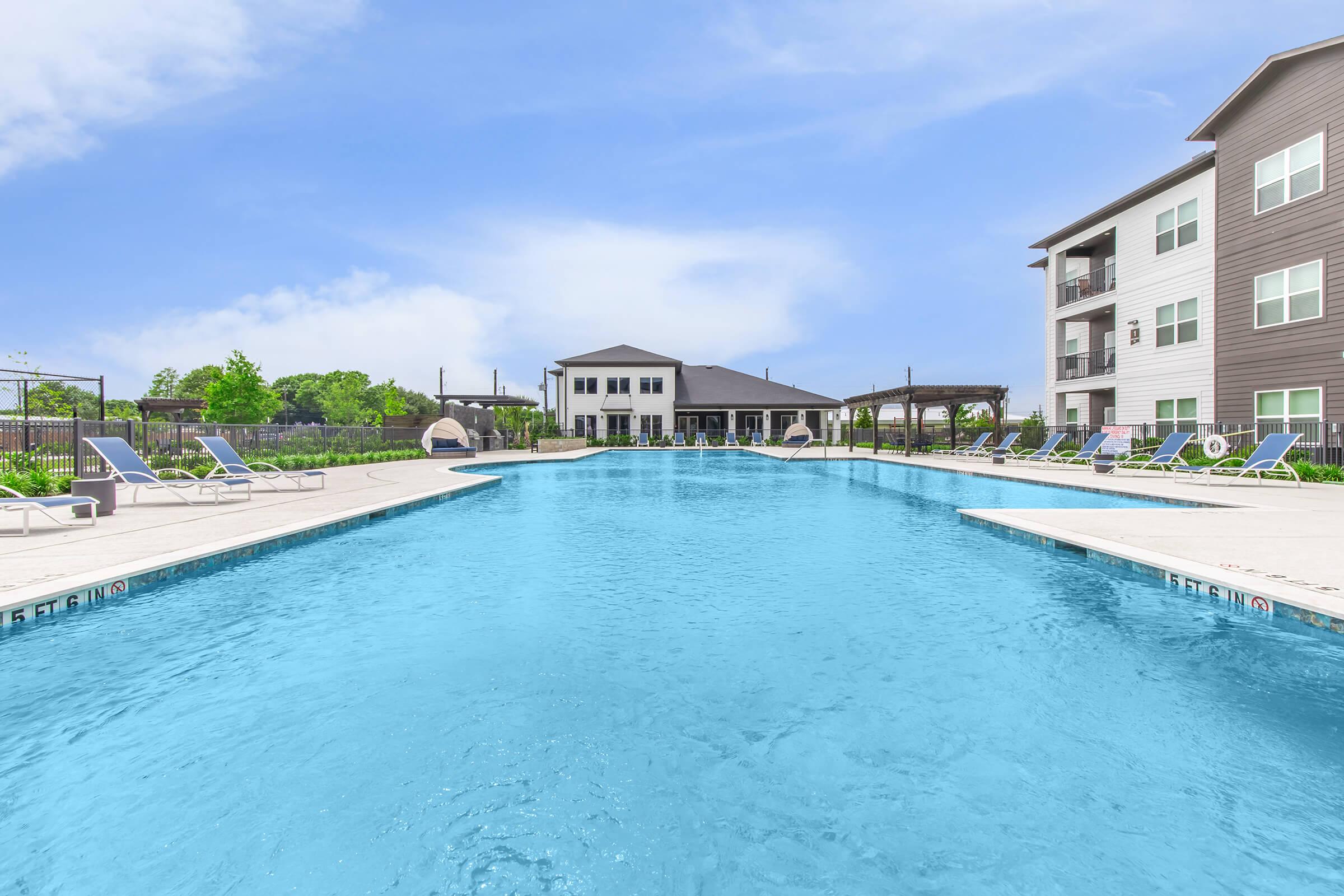 A large, inviting swimming pool with crystal-clear water is surrounded by lounge chairs. In the background, there is a modern building and a covered patio area. Lush greenery and a clear blue sky complete the serene outdoor setting.