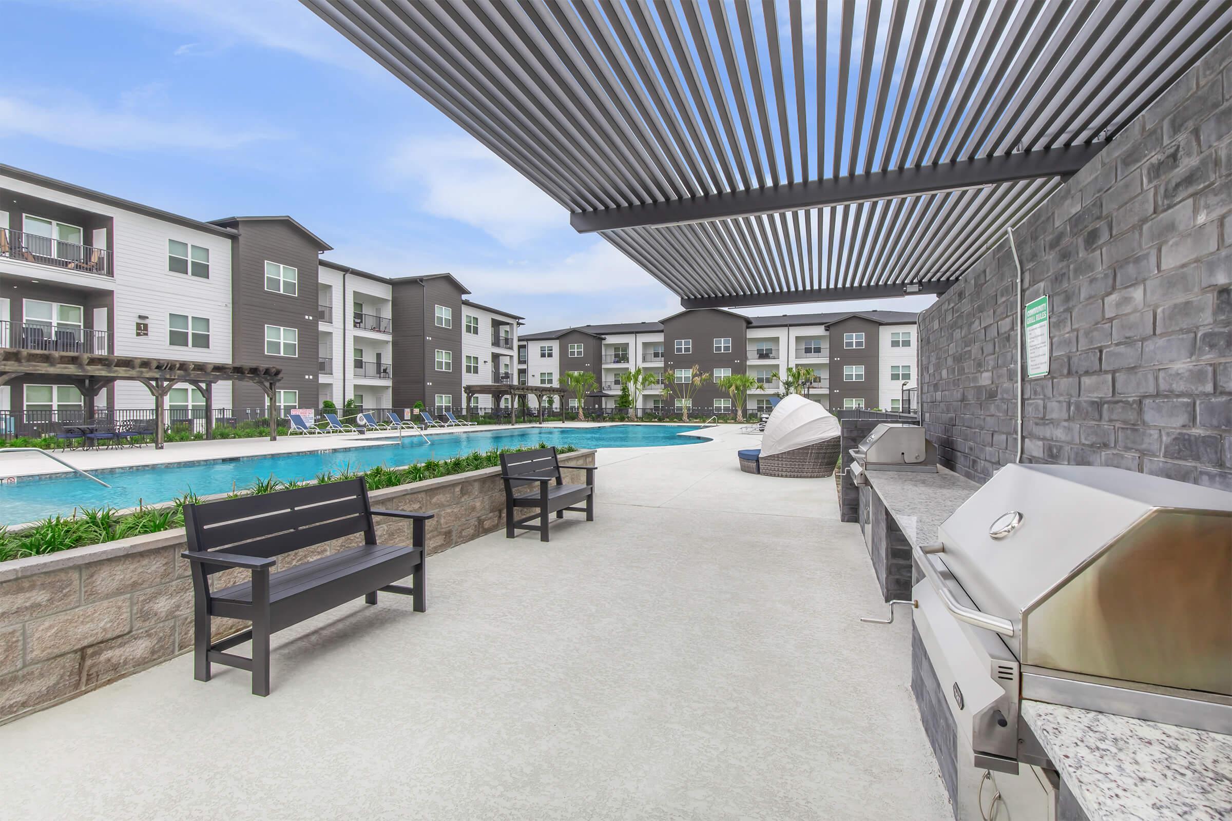 A modern apartment complex with a swimming pool. In the foreground, there are two benches and a barbecue grill under a shaded area. The pool, surrounded by lounge chairs, is visible in the background, with well-maintained landscaping and contemporary buildings lining the far side.