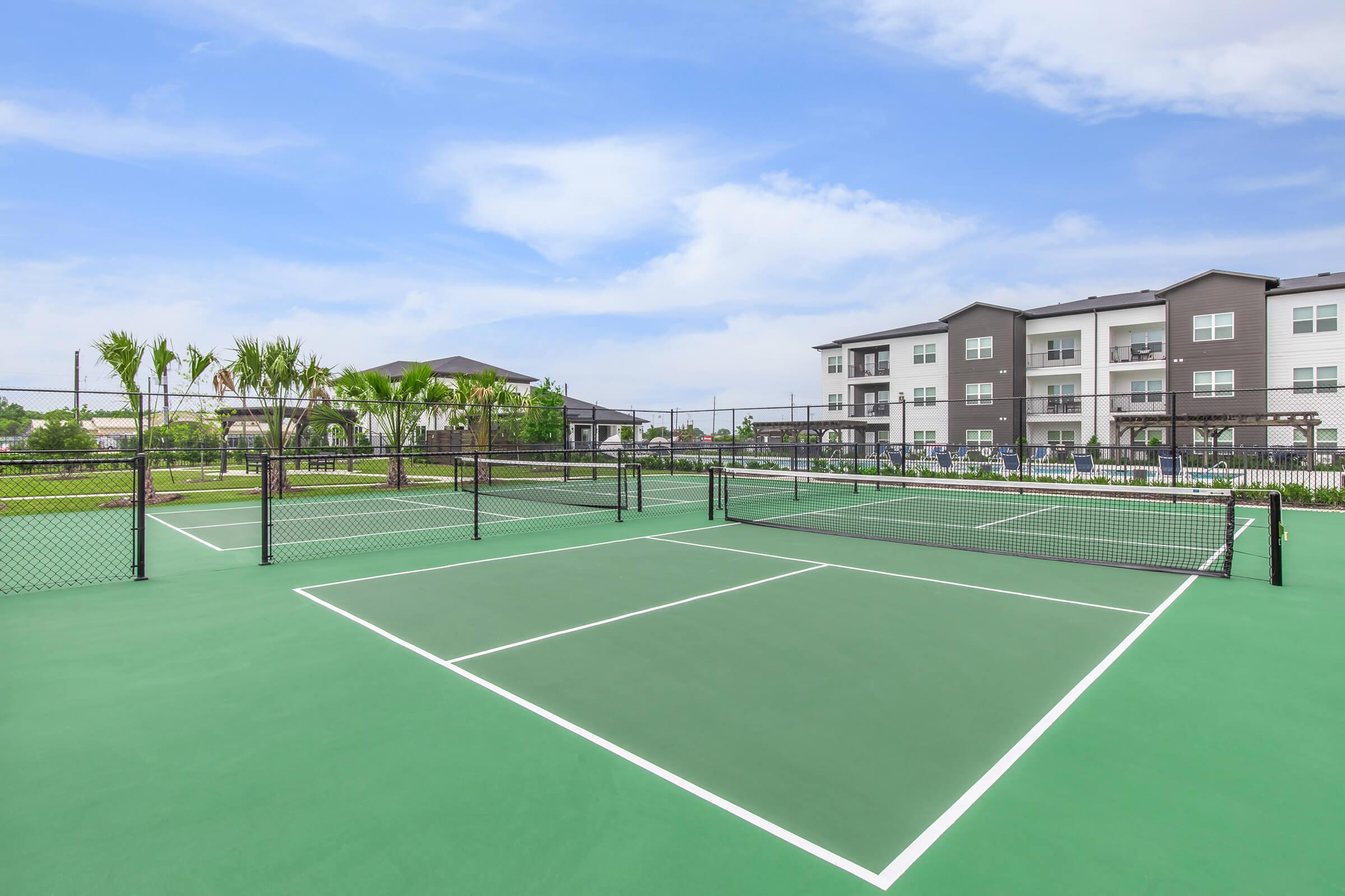 Tennis courts with green surfaces and black fencing, surrounded by palm trees and modern residential buildings in the background. The sky is partly cloudy, and the area appears well-maintained and inviting for recreational activities.