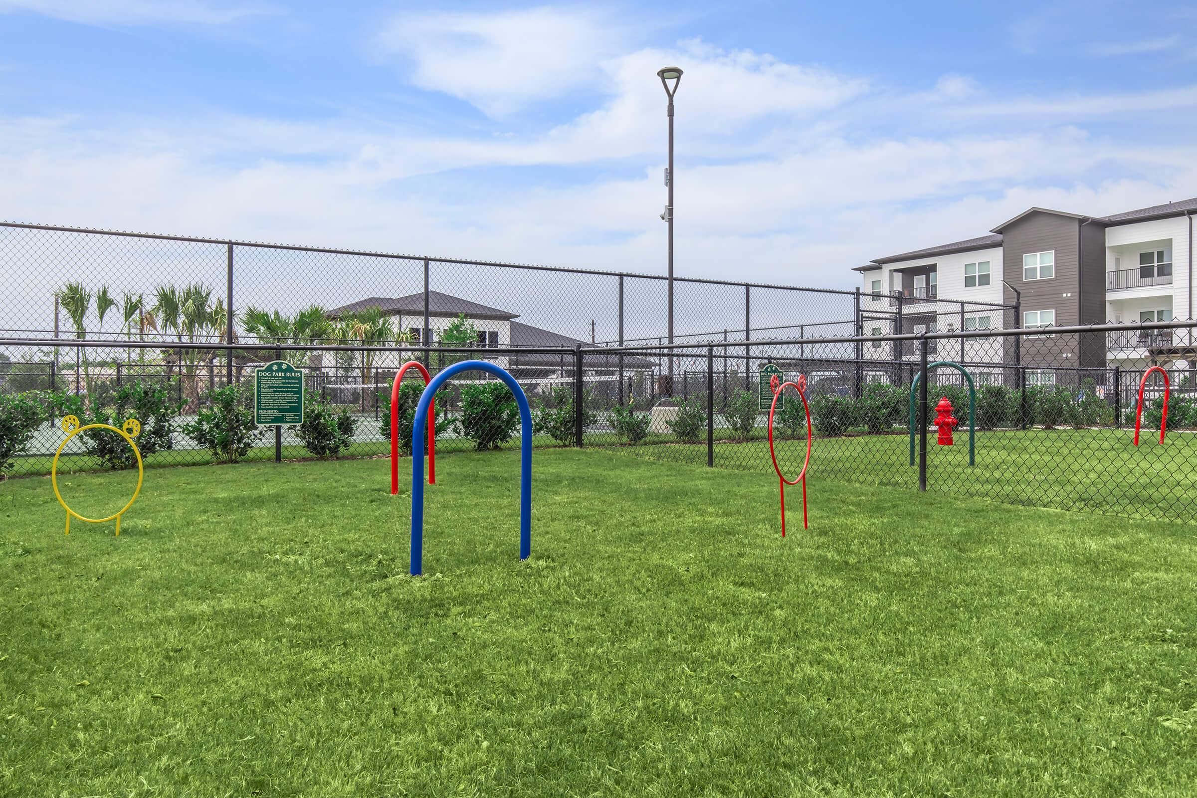 A fenced dog park featuring colorful agility equipment, including hoops in various colors, set on a grassy area. The background includes a modern apartment building and palm trees, with a clear blue sky overhead.