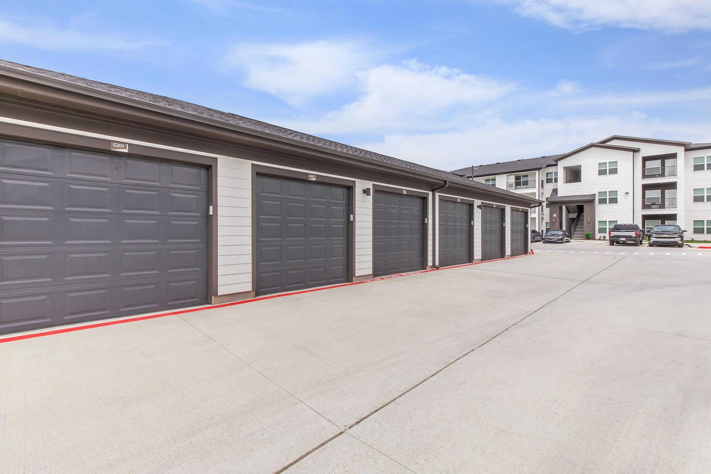 A row of closed garage doors in a modern apartment complex. The garages are located on a paved surface with a clear blue sky in the background. Nearby, several parked cars are visible in front of a multi-story building.