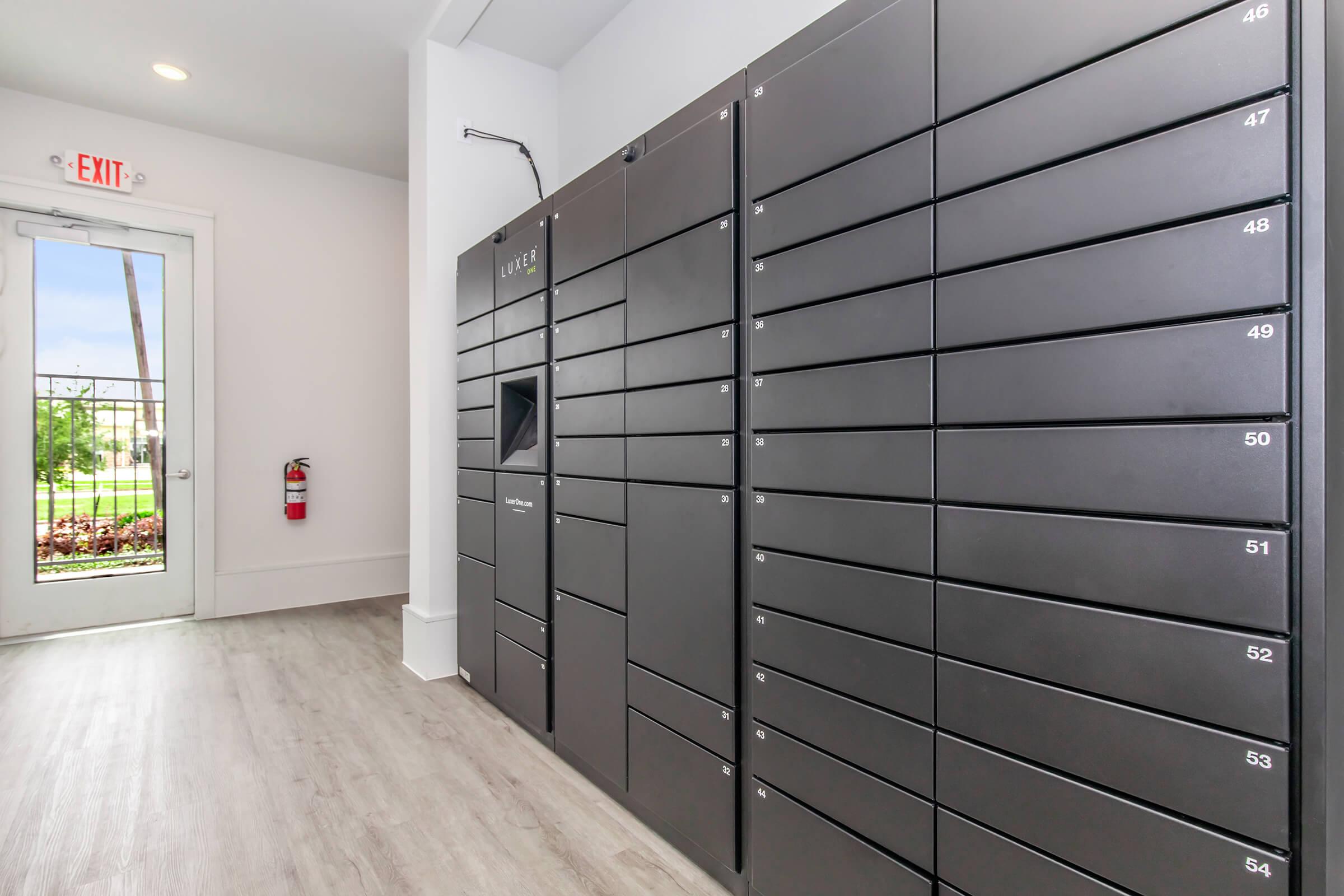 Rows of metallic gray delivery lockers line a wall in a modern interior space. The lockers are numbered from 46 to 56, with one larger locker in the center. A door leads outside, and a fire extinguisher is visible nearby. Natural light enters through the door, illuminating the area.