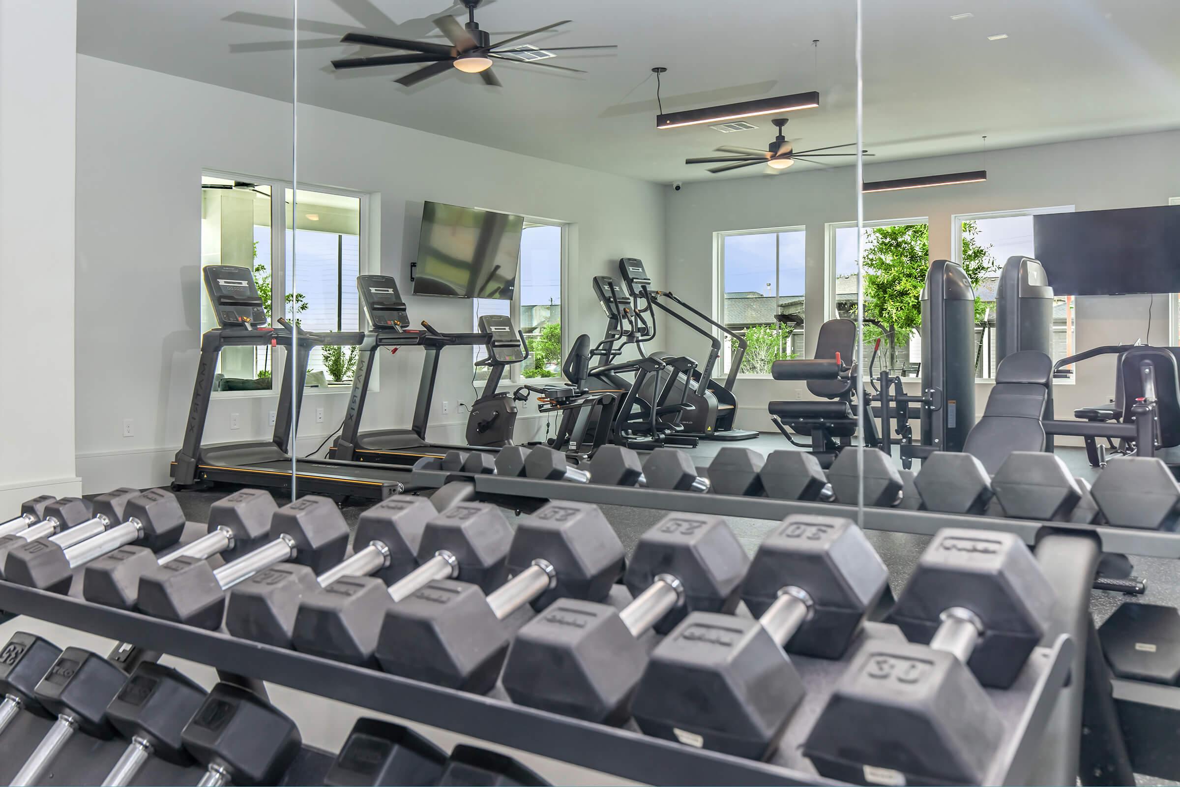 A modern gym interior featuring a rack of dumbbells in the foreground. Reflective surfaces show various cardio machines like treadmills and ellipticals in the background, along with large windows letting in natural light. The space is equipped with multiple screens and has a clean, organized appearance.
