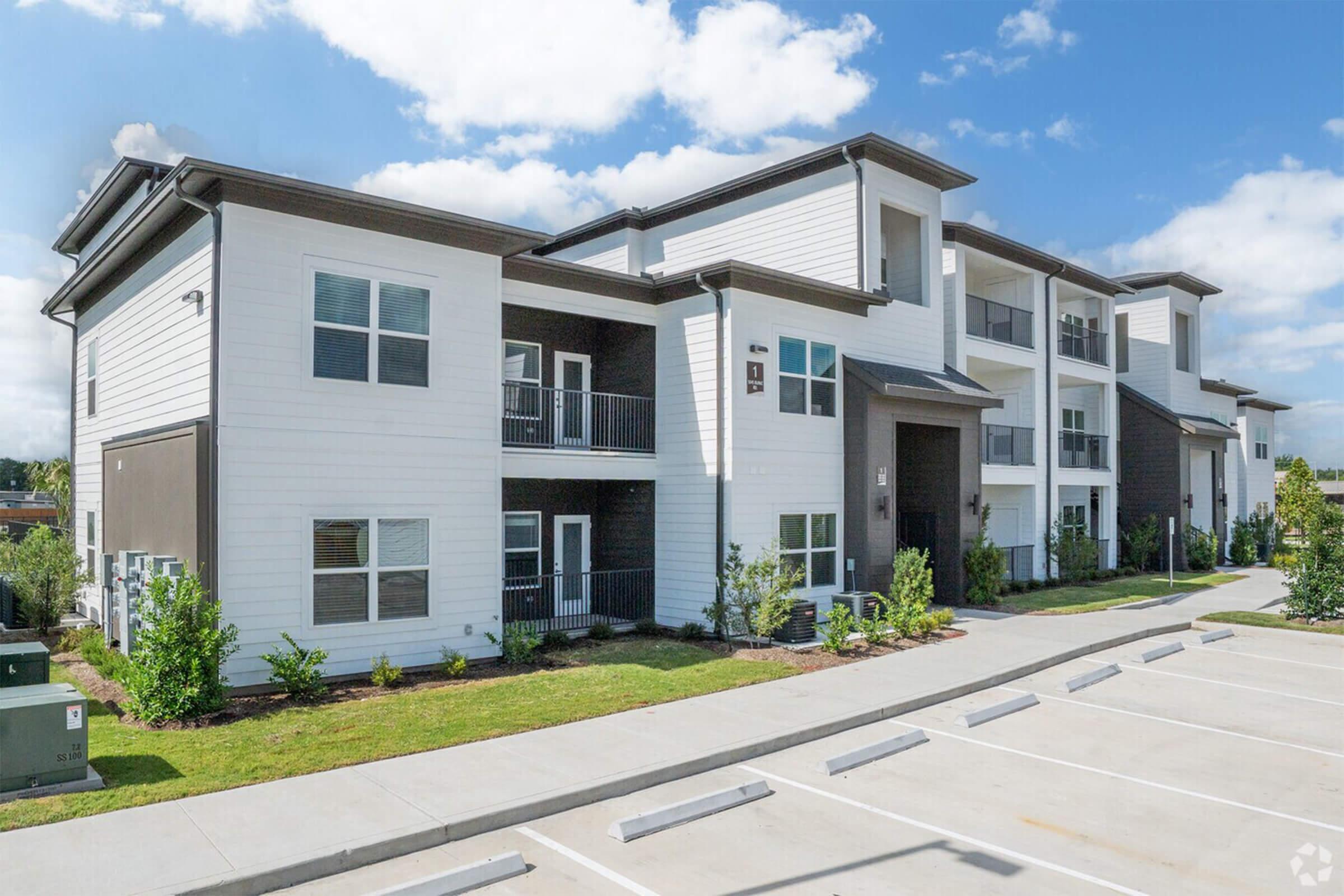 Modern apartment complex with three stories and a mix of white and dark exterior finishes. The building features balconies on the upper floors and landscaped grounds. A parking lot with multiple spaces is visible in the foreground, set against a clear blue sky.