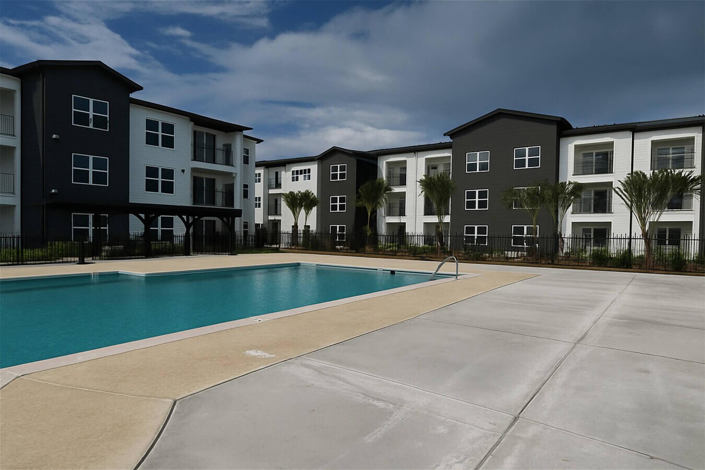 A modern apartment complex with two buildings, featuring a swimming pool in the foreground. The pool is surrounded by a patio area and palm trees, under a cloudy sky. The buildings are painted in a mix of white and dark gray, with multiple windows.