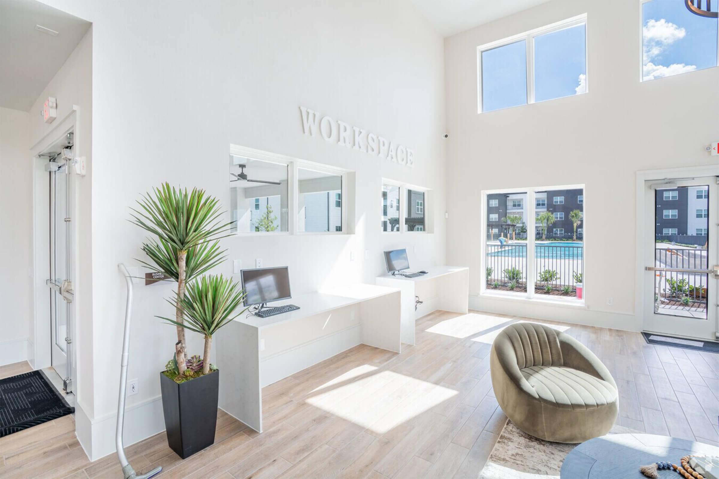 Bright and airy workspace featuring two computer stations and a green plant. Large windows allow natural light to flood in, overlooking a swimming pool area. The modern decor includes a cozy green armchair and sleek countertops, creating a welcoming environment for productivity.