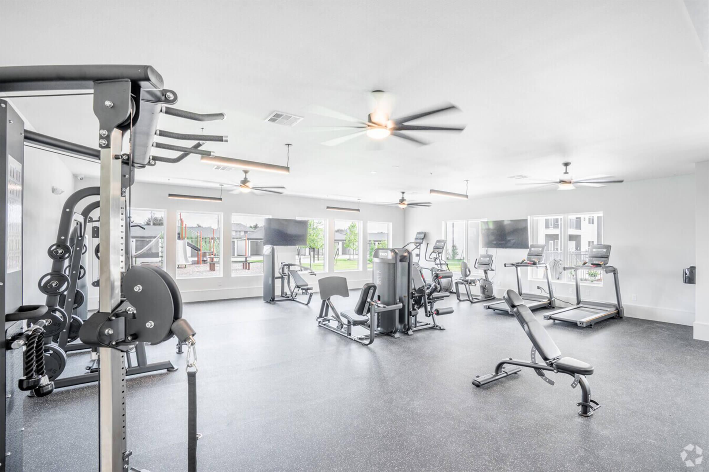 A modern residential fitness center featuring various gym equipment, including weight machines, treadmills, and stationary bikes. Bright, spacious with large windows allowing natural light, and ceiling fans for ventilation. The room is designed for workouts and physical fitness activities.