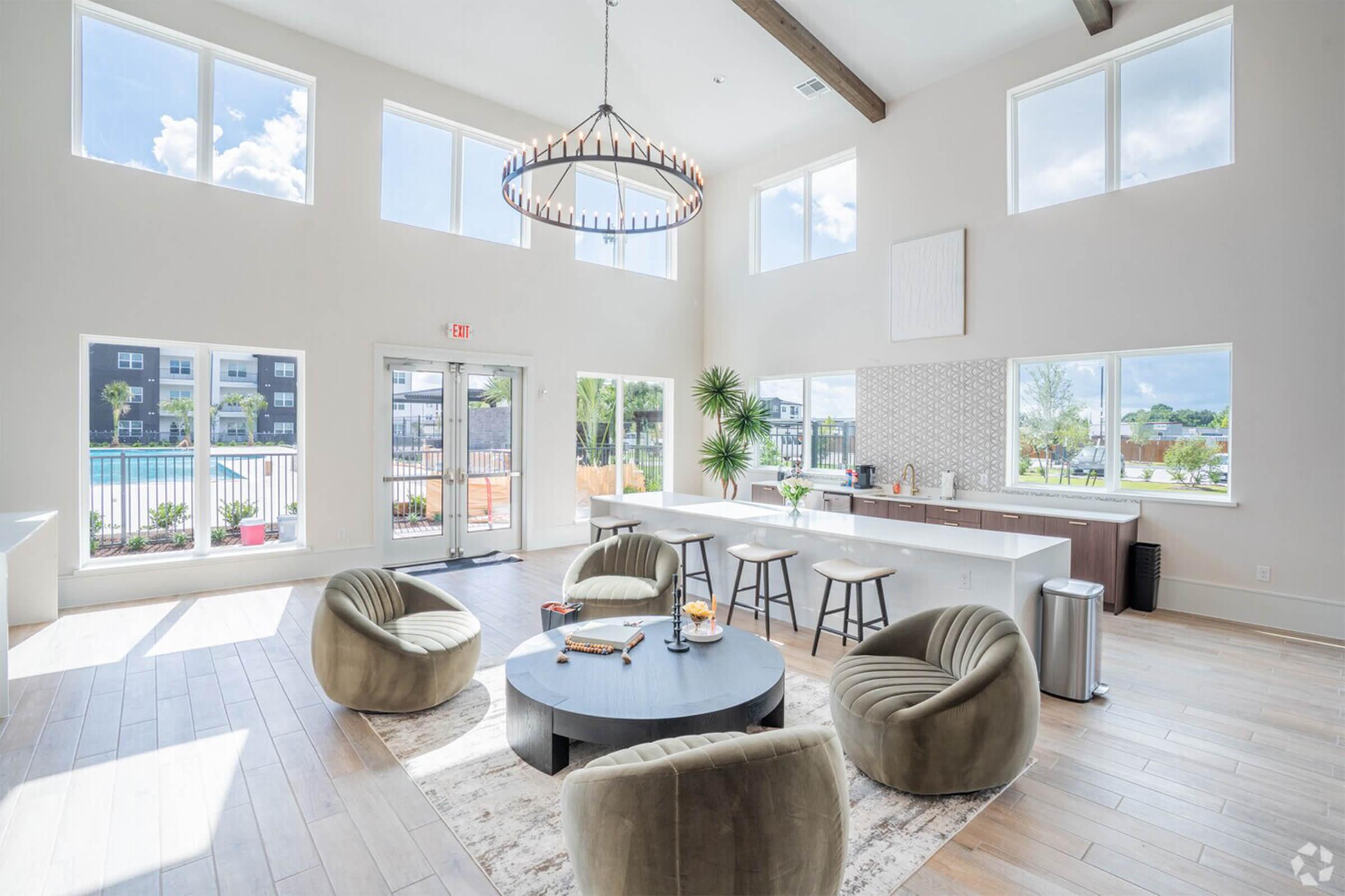 A bright, modern lounge area featuring large windows, a central circular table with snacks, and plush chairs. In the background, a kitchenette with bar seating and a view of a pool area. The high ceiling and light colors create an inviting atmosphere.