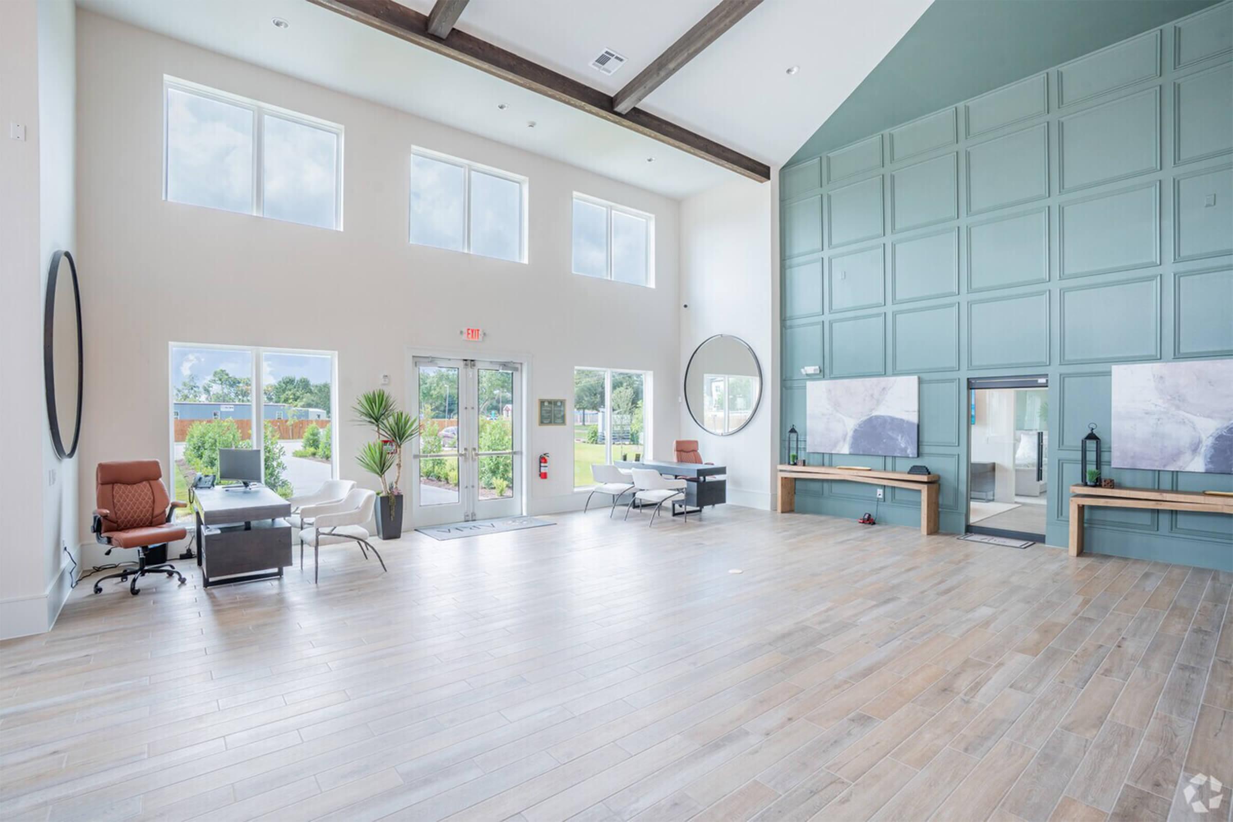 A spacious lobby area featuring large windows, wooden flooring, and modern furnishings. There are two seating areas with white chairs, a desk with a brown chair, and decorative plants. A large round mirror and framed artwork adorn the walls, creating a welcoming atmosphere with ample natural light.