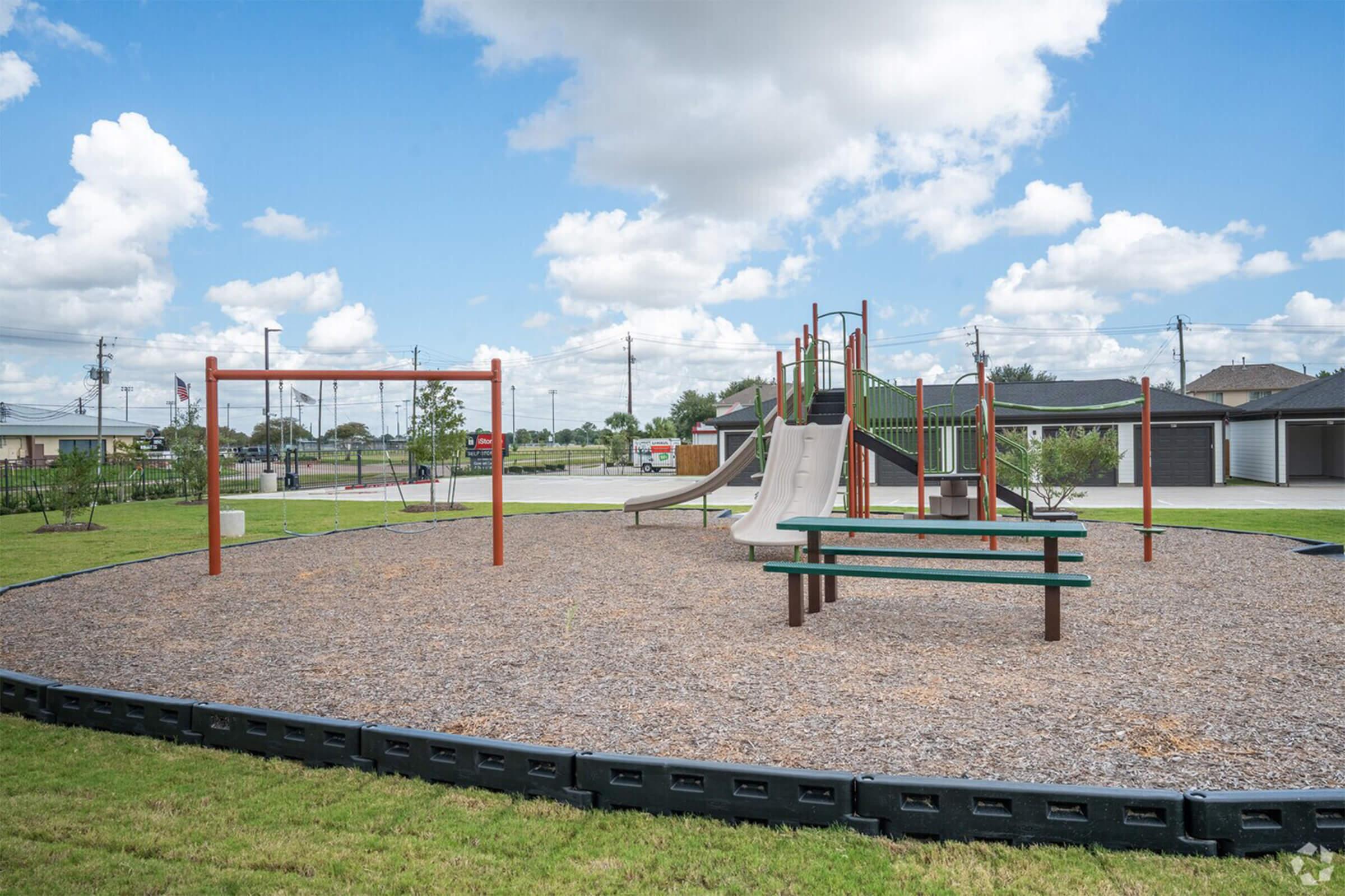 A playground scene featuring wooden swings, a slide, and a climbing structure, surrounded by a gravel area. There is a bench for seating, and the background shows grassy fields and a blue sky with fluffy clouds. Utility poles and buildings are faintly visible in the distance.