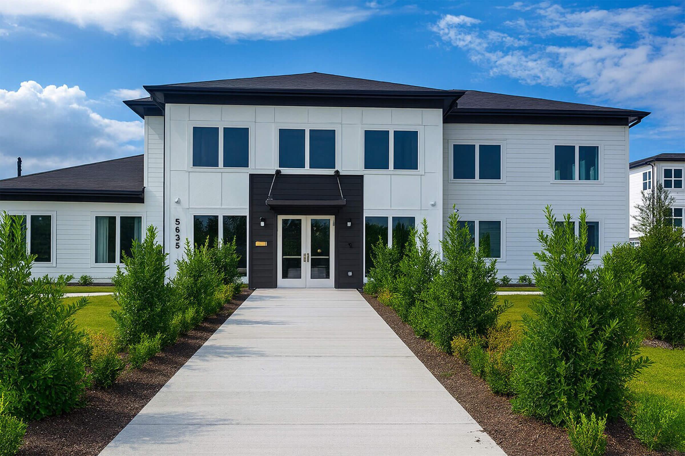 A modern two-story building featuring a combination of white and dark siding. It has multiple large windows and a welcoming entrance. A well-maintained pathway flanked by young shrubs leads up to the front door, set against a backdrop of a blue sky with scattered clouds.
