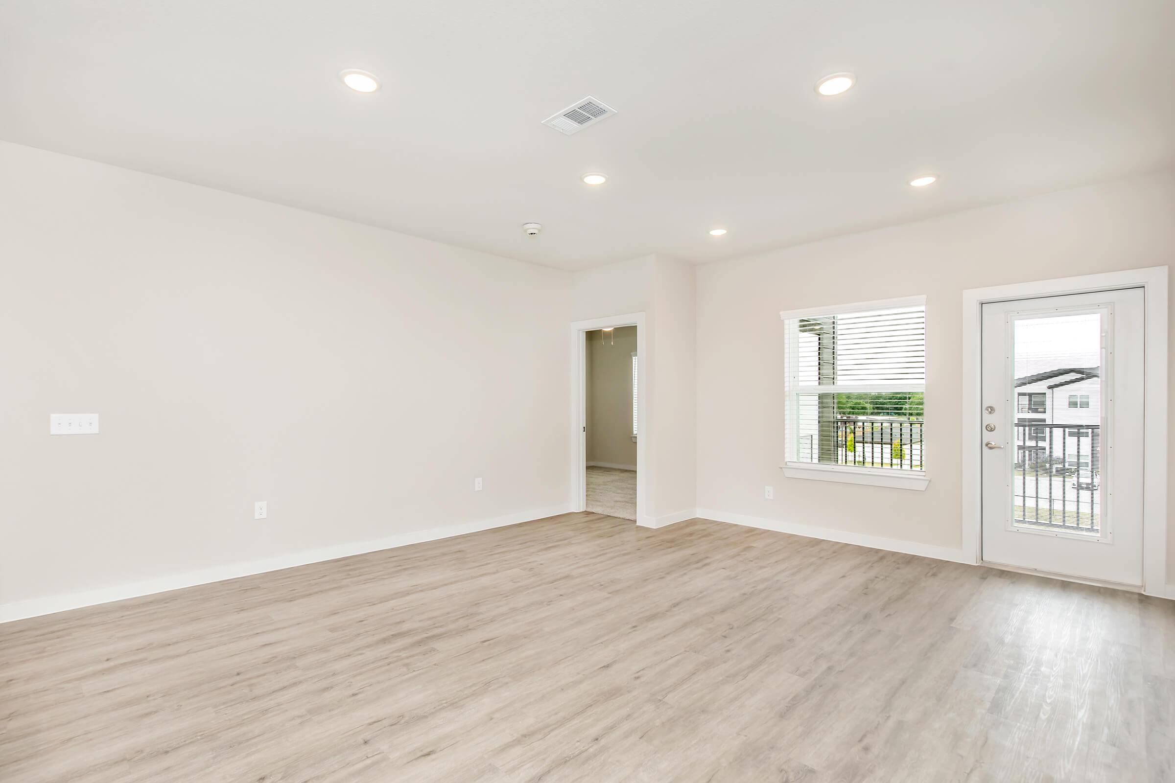 A spacious, well-lit living room featuring light-colored walls and hardwood flooring. There are recessed ceiling lights and a large window with white blinds. A doorway leads to another room, and a glass door opens to a small balcony or patio area. The overall atmosphere is modern and inviting.