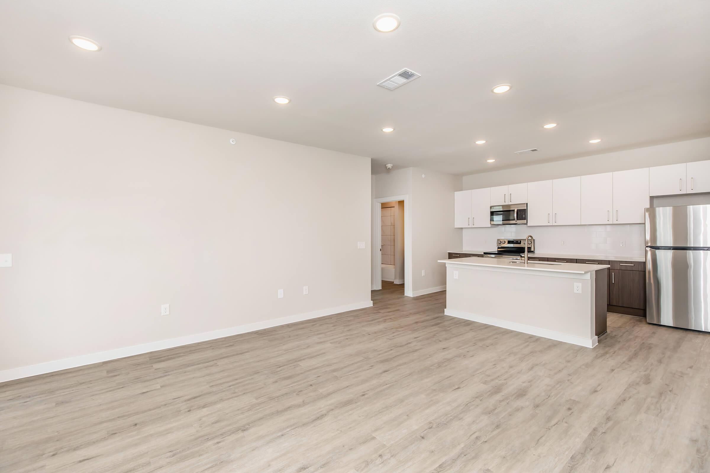 A modern, open-concept kitchen and living area featuring light-colored walls, wood-like laminate flooring, recessed lighting, and stainless steel appliances. The kitchen includes a countertop bar with seating and is designed for a spacious and inviting atmosphere.