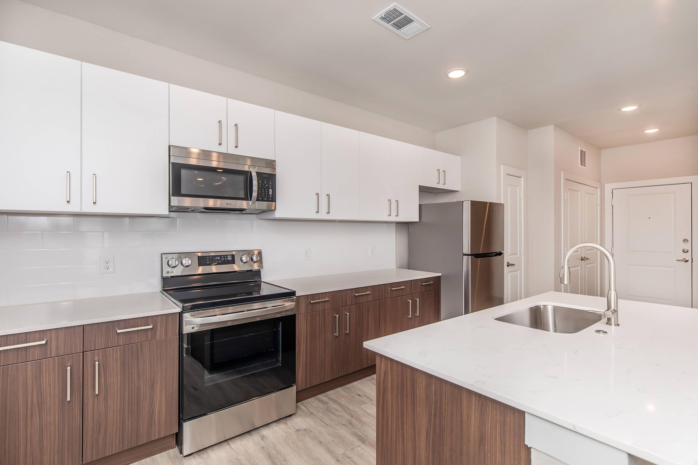 Modern kitchen featuring stainless steel appliances, including a microwave and oven, with sleek white cabinetry and a contrasting dark wood island. The countertop is made of light-colored stone, and there's a single-basin sink with a modern faucet. The space is well-lit with recessed lighting.