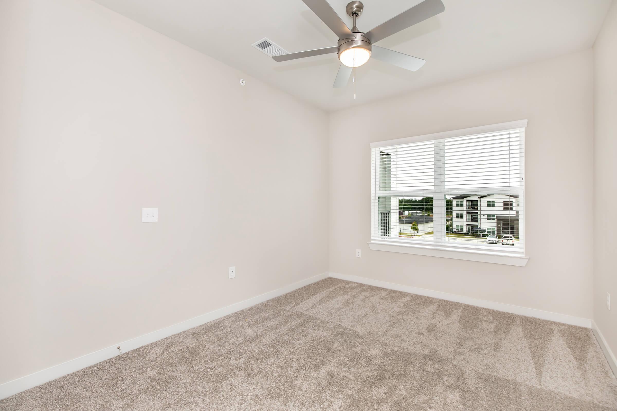 A vacant room with light beige walls and carpet. There is a ceiling fan and a large window with white blinds, revealing an outside view. The room has no furniture, creating a bright and spacious atmosphere.