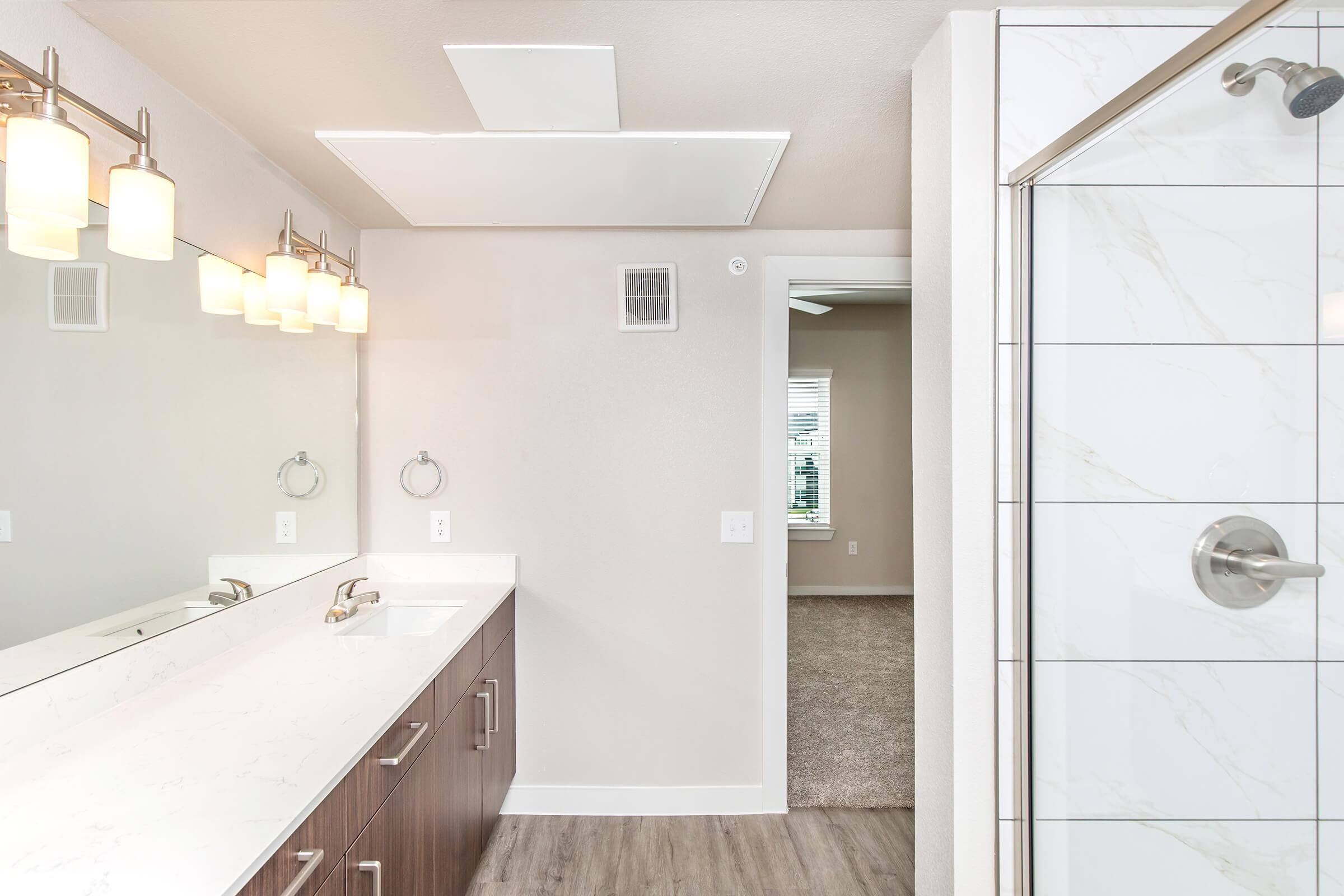 A modern bathroom featuring a double vanity with a white quartz countertop, two wall-mounted round mirrors, and stylish light fixtures. A glass shower enclosure with marble-like tiles and a nearby door leading to a carpeted room can be seen in the background. The walls are painted in a light, neutral color.