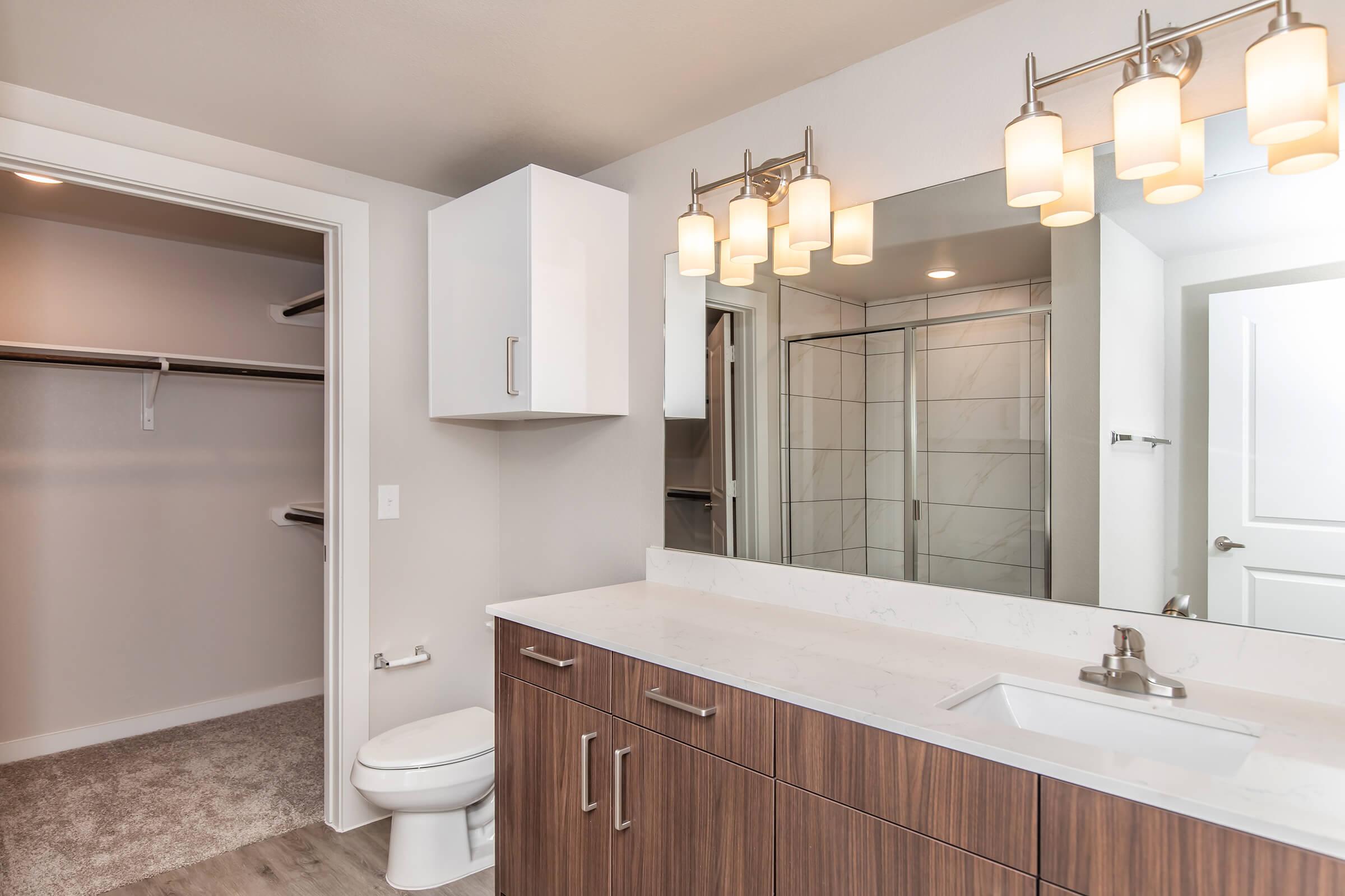 A modern bathroom featuring a light-colored vanity with a white countertop and multiple storage cabinets. There is a large mirror above the sink, accompanied by stylish lighting fixtures. A glass shower is visible, alongside a toilet and a spacious closet with hanging space. The walls are painted in a soft neutral tone.