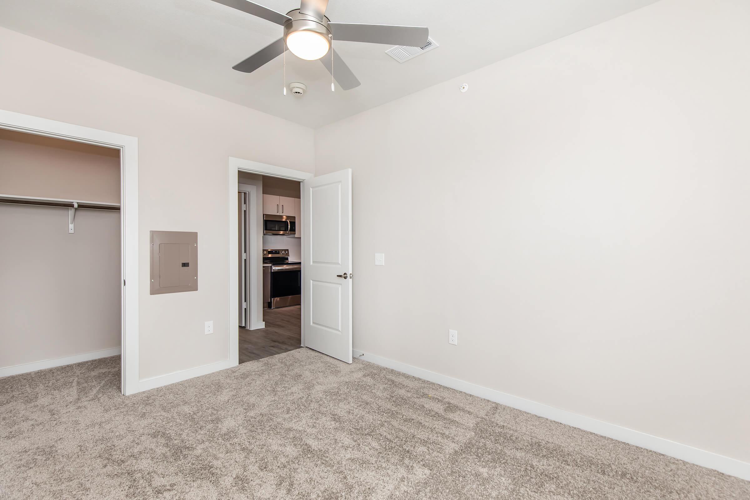 A well-lit, empty room featuring a ceiling fan, light beige walls, and plush carpet. The room includes a white closet with sliding doors on the left and an open door leading to another space. The overall aesthetic is modern and minimalistic, creating a spacious feel.