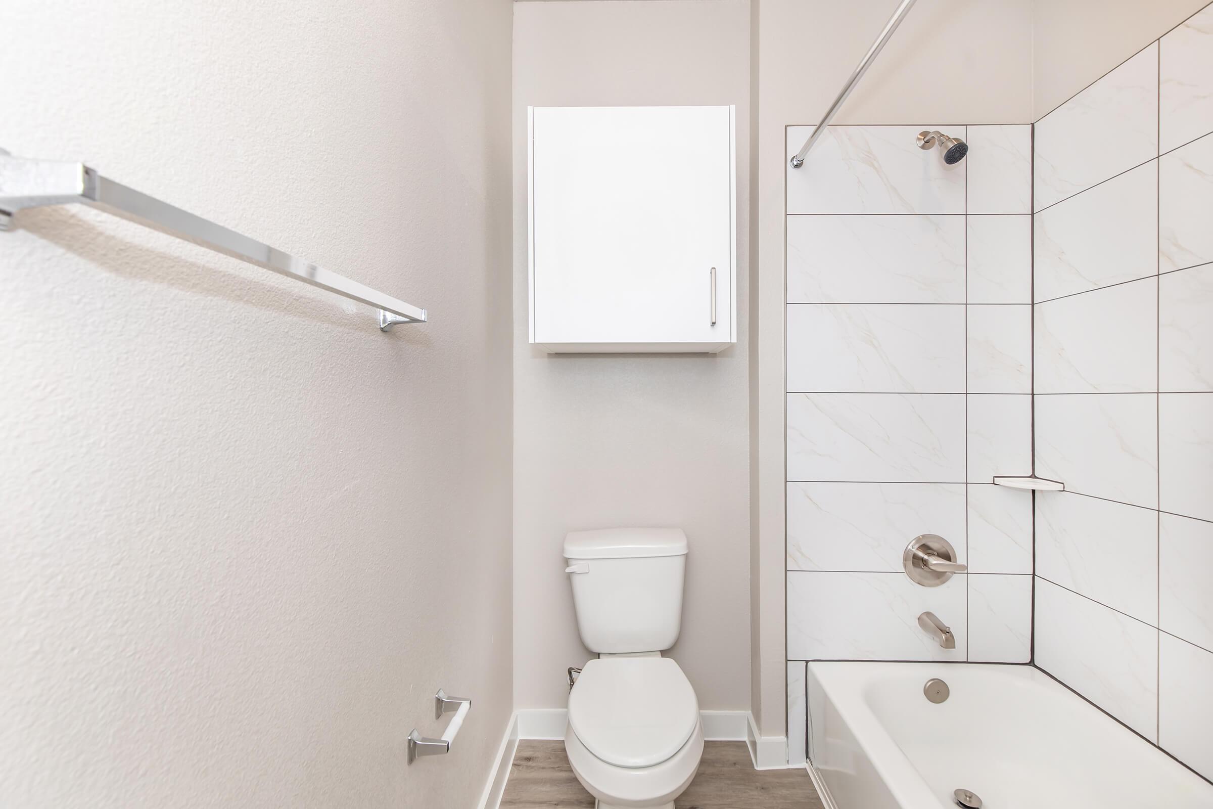 A clean, modern bathroom featuring a white toilet, a bathtub with a showerhead, and a wall-mounted cabinet above. The walls are finished in light colors, with sleek tile accents in the shower area. A stainless steel towel bar is mounted on the wall, contributing to the minimalist design.