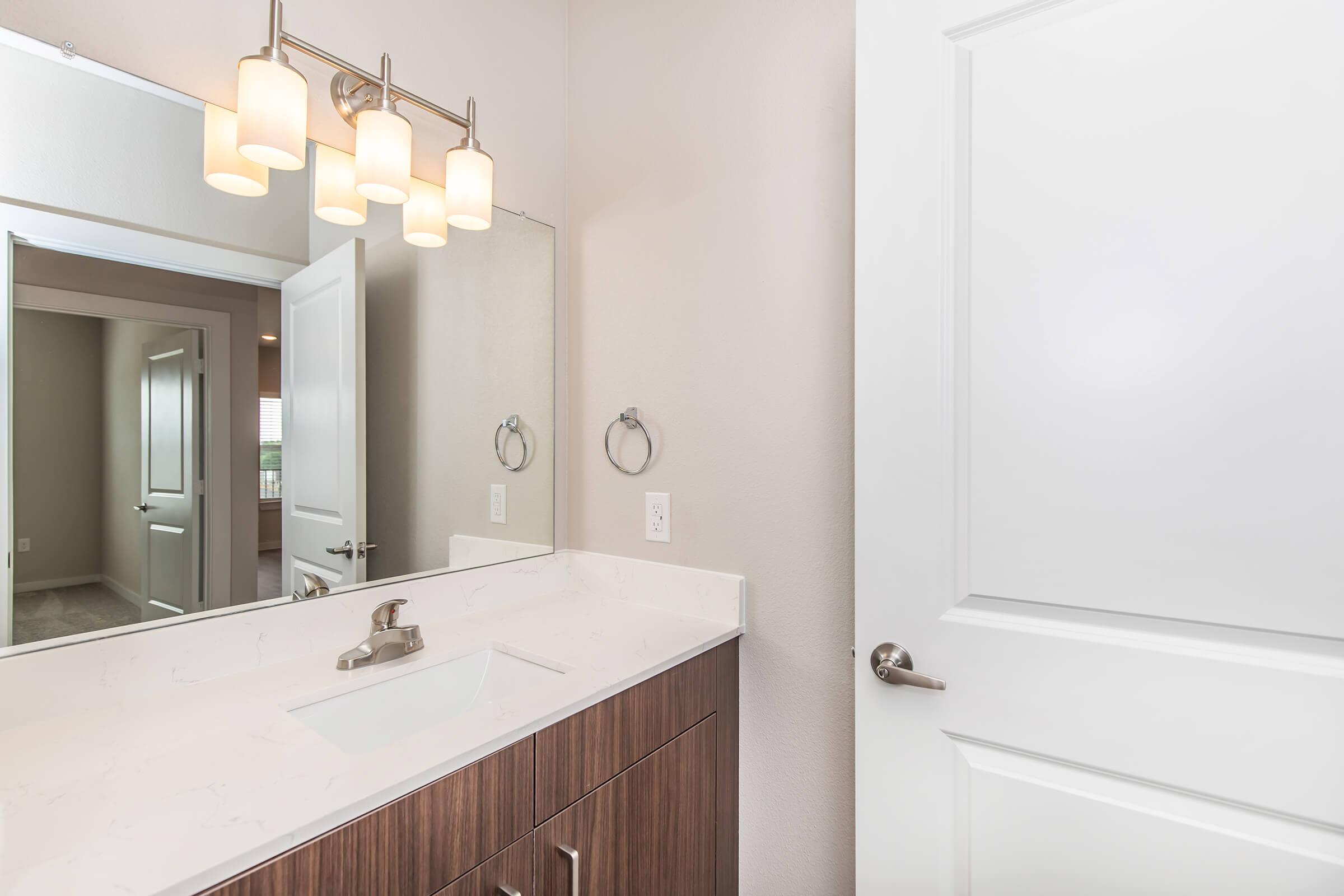 A modern bathroom featuring a light-colored wall, a white countertop with an under-mount sink, and a stylish faucet. Above the sink, three pendant lights provide illumination. A mirror reflects the space, and a door is visible in the background, leading to another room. The cabinetry has a wood finish.