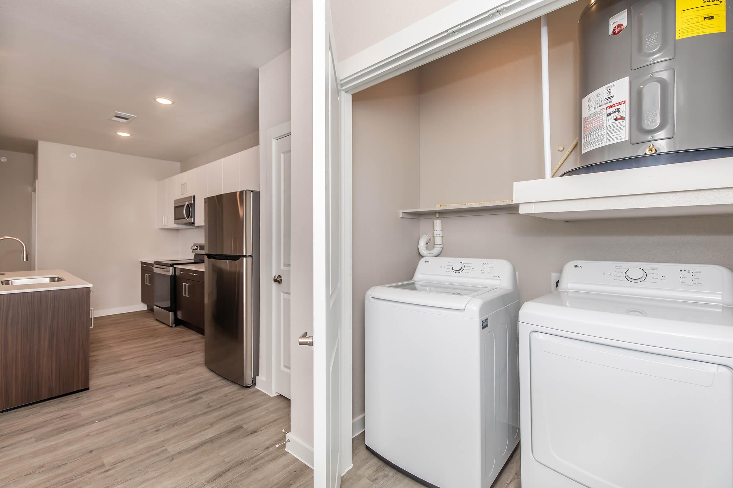 A modern kitchen and laundry area featuring stainless steel appliances, including a refrigerator, microwave, and oven. The laundry area has white washing machines and a dryer, with a shelf above. The flooring is a light wood look, creating a clean and contemporary aesthetic.