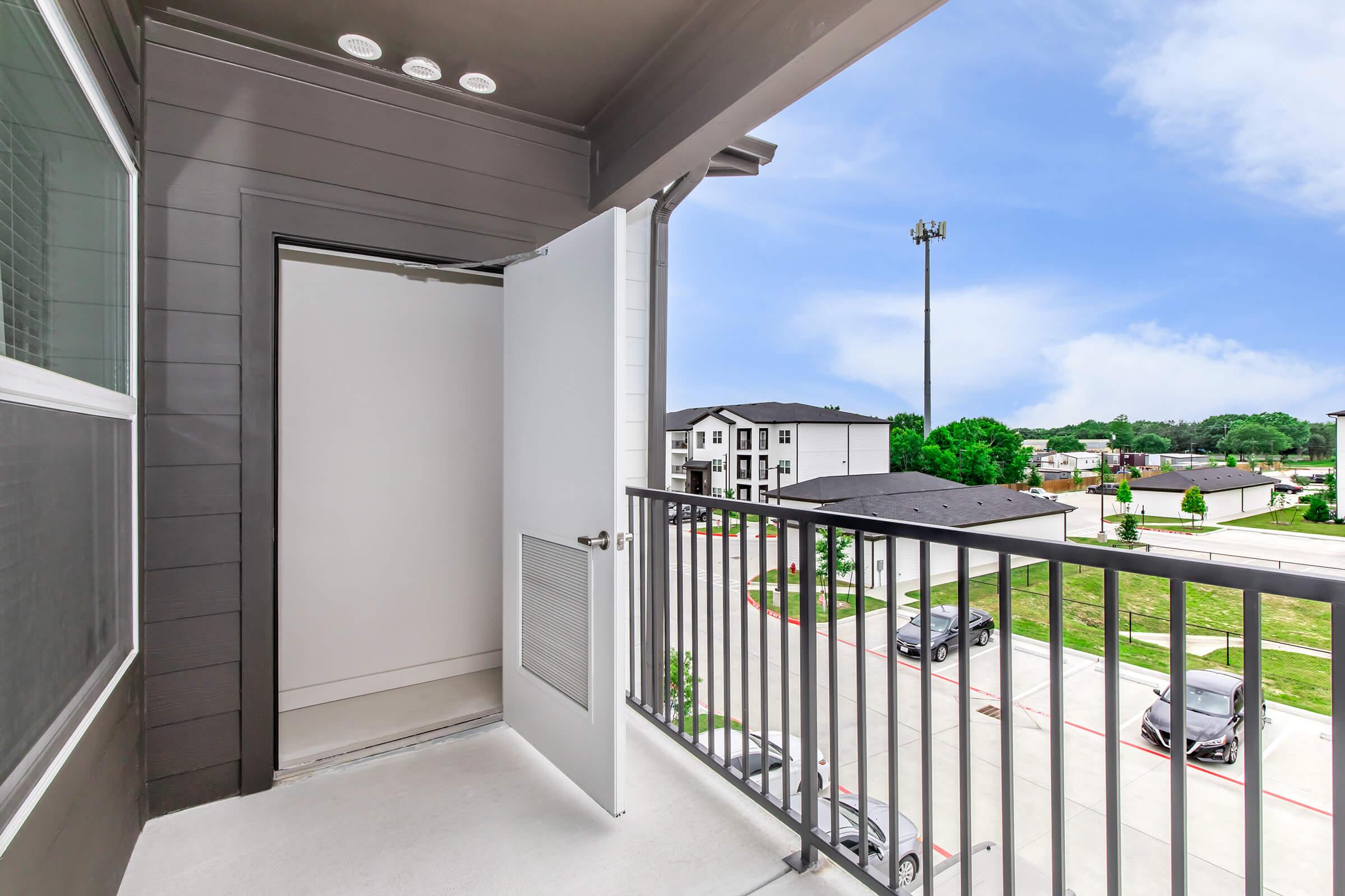 A balcony view featuring a white door leading into a room, metal railing, and a landscaped area with parked cars below. In the background, there are modern apartment buildings and a cell tower, with a clear blue sky overhead.
