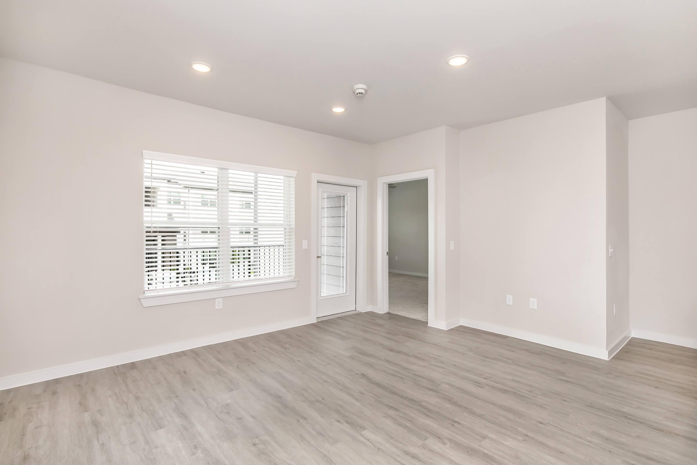 A spacious, empty room with light-colored walls and wood-like flooring. Large windows with white blinds allow natural light to enter. A door leads to an outdoor area, while an adjacent doorway opens to another room. The overall atmosphere is bright and airy, suitable for various interior design styles.