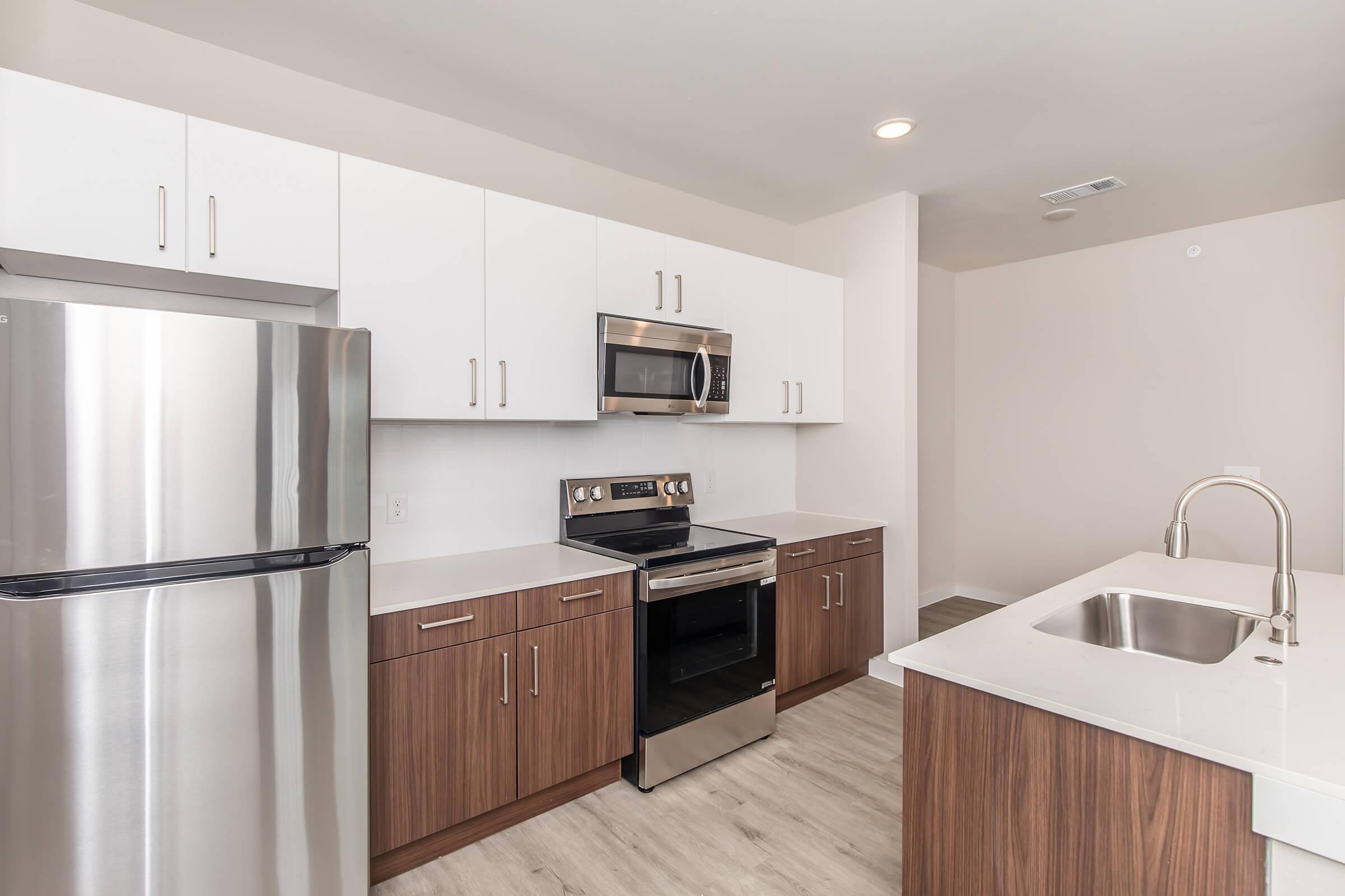 Modern kitchen featuring stainless steel appliances including a refrigerator, microwave, and oven. The cabinetry is a mix of white and wood tones, with a sleek countertop and a large stainless steel sink. The flooring is light-colored, enhancing the spacious feel of the kitchen.