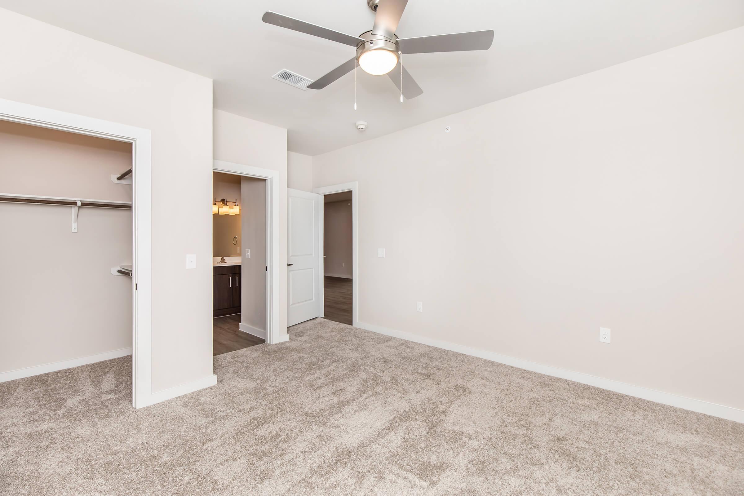 A spacious, empty room with light-colored walls and beige carpet. Features include a ceiling fan, a closet with open doors to the left, and two doorways leading to other areas. Natural light fills the room, highlighting the clean and modern design.
