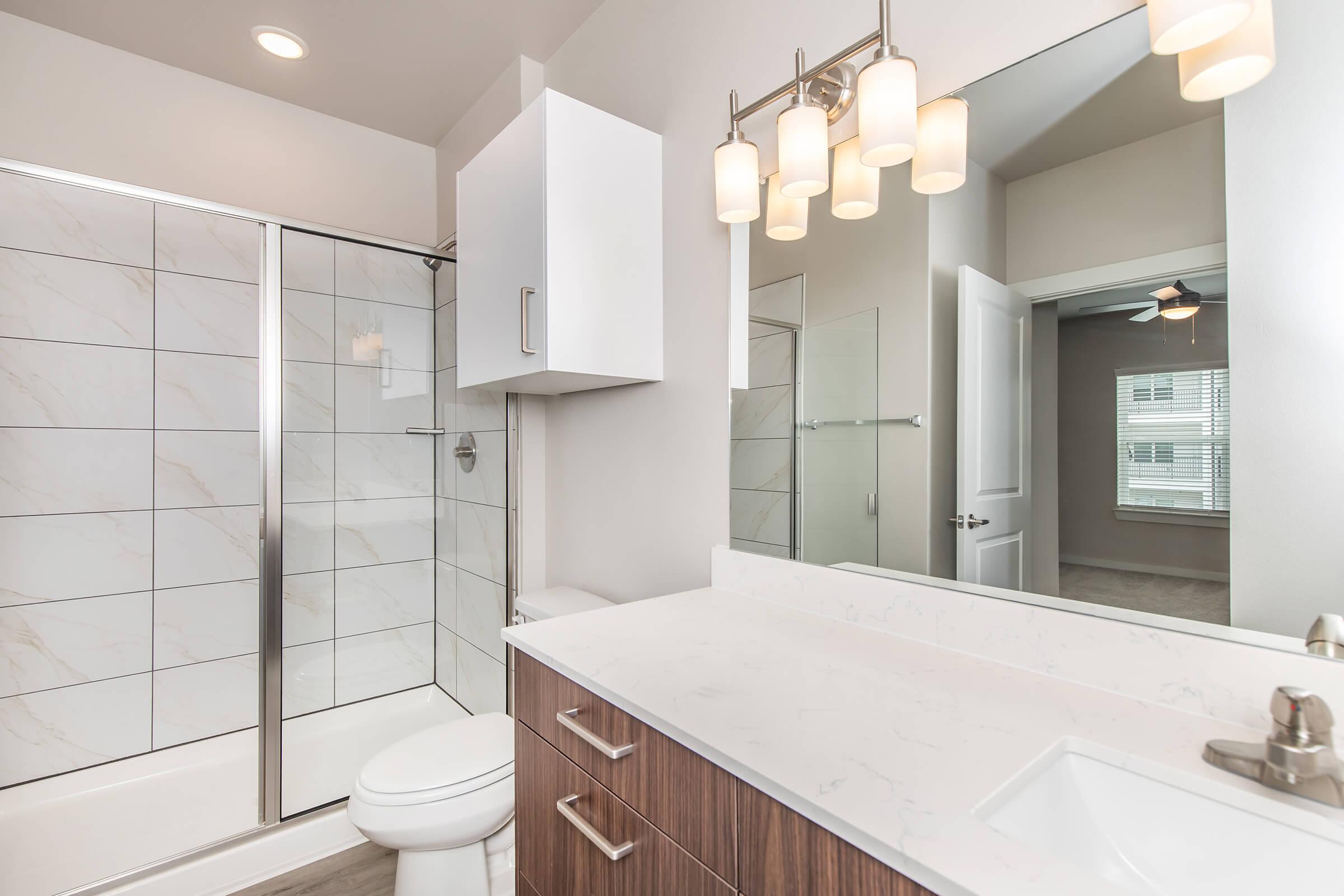 Modern bathroom featuring a glass shower enclosure, a white bathtub, and a sleek vanity with dark wood cabinetry. A large mirror is above the sink, illuminated by stylish pendant lights. The room has neutral-colored walls and a window allowing natural light, adding to the spacious feel.