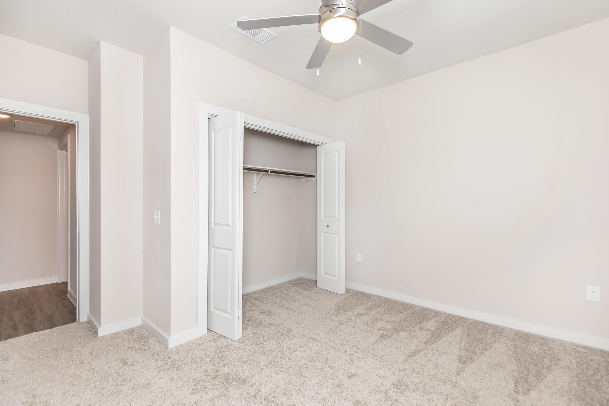 A well-lit, empty room featuring beige walls, a ceiling fan, and a carpeted floor. On one side, there are two open white doors leading to a closet with a hanging rod. The room appears spacious, with a clear view of adjacent walls and no furniture present.