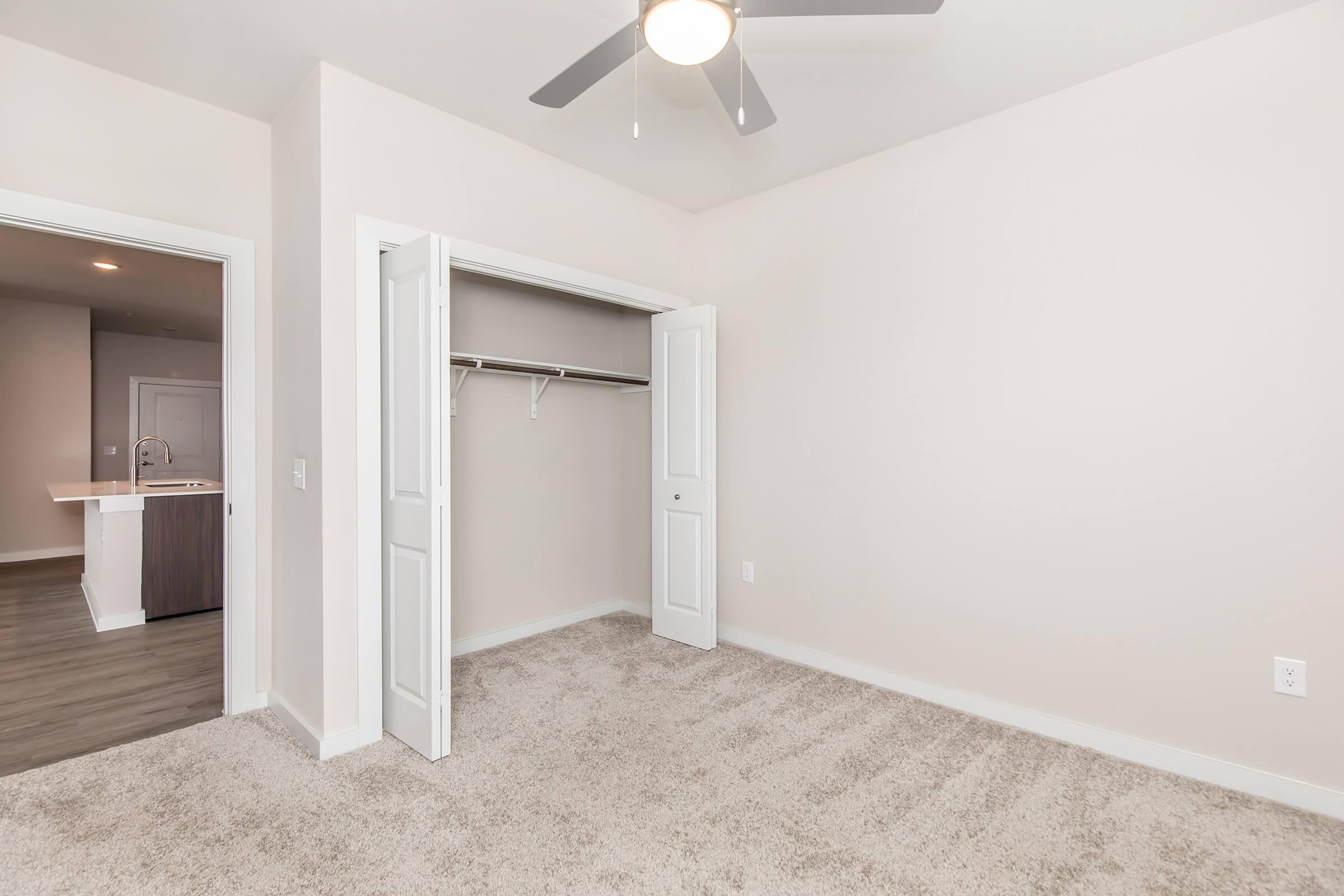 Empty room featuring light-colored walls and carpet. There is a spacious closet with sliding doors on one side. A ceiling fan is mounted above, and the entrance to another room is visible in the background, along with a glimpse of a modern kitchen area. Natural light illuminates the space.