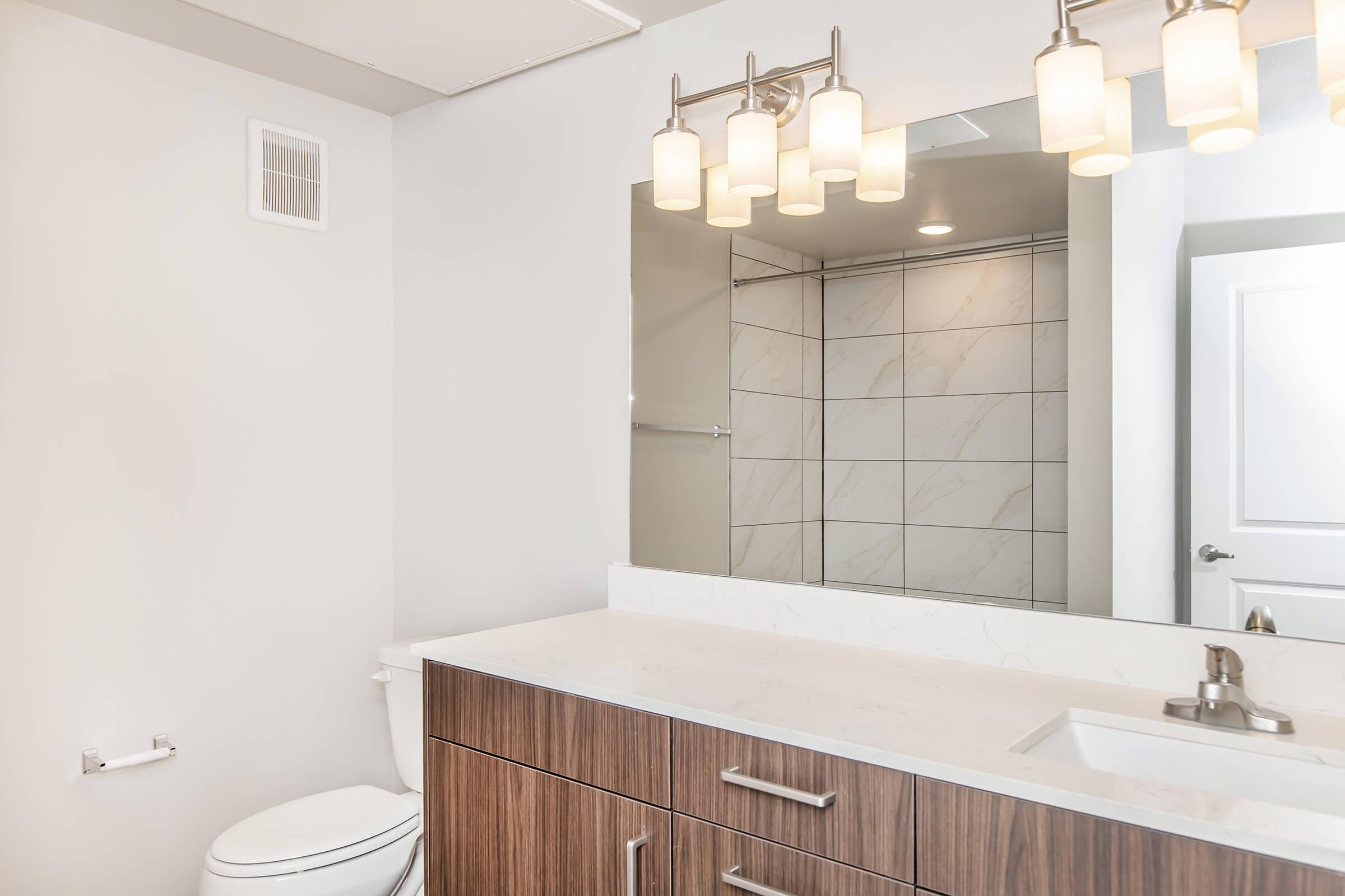 A modern bathroom featuring a wooden vanity with a white countertop and sink, a large mirror, illuminated by stylish light fixtures. The wall is tiled with a marble pattern, and the space includes a toilet and a towel holder. Natural light brightens the clean, minimalist decor.