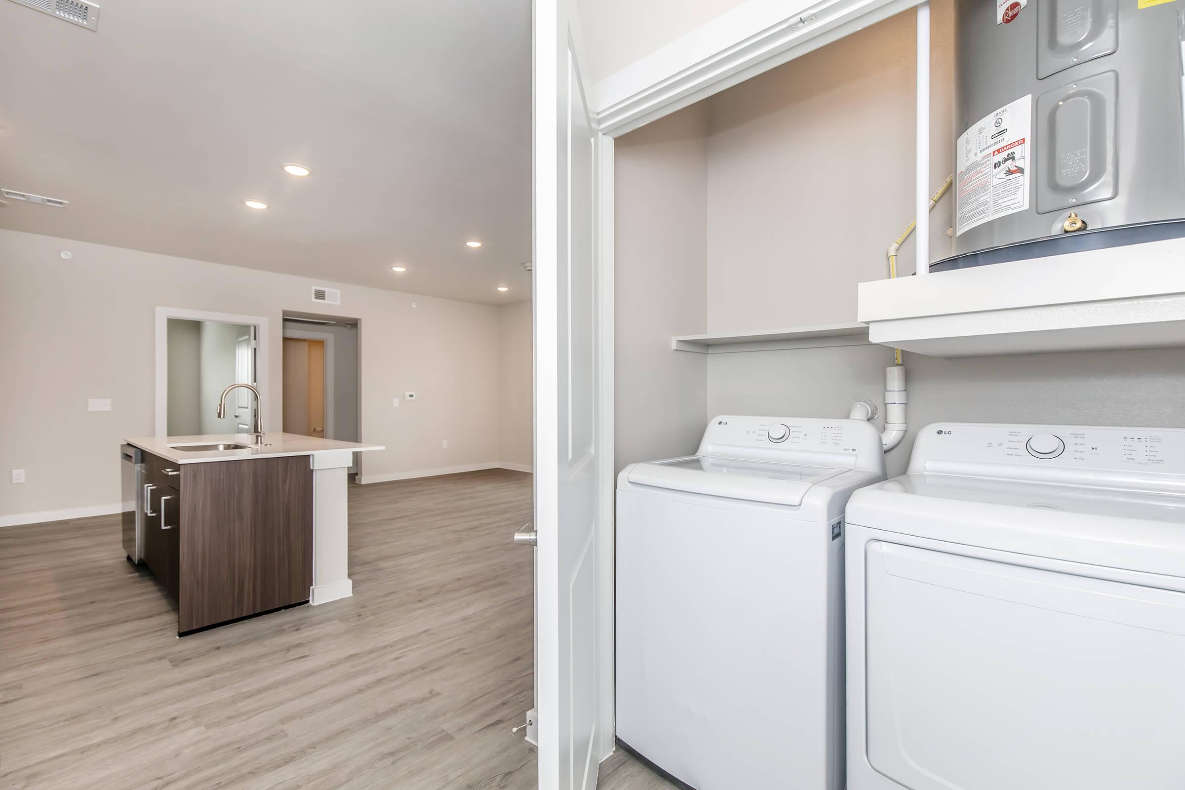 A view from a laundry area showing two white washing machines side by side, with a water heater on a shelf above. The space leads into an open concept kitchen and living area with wood-like flooring and modern lighting.